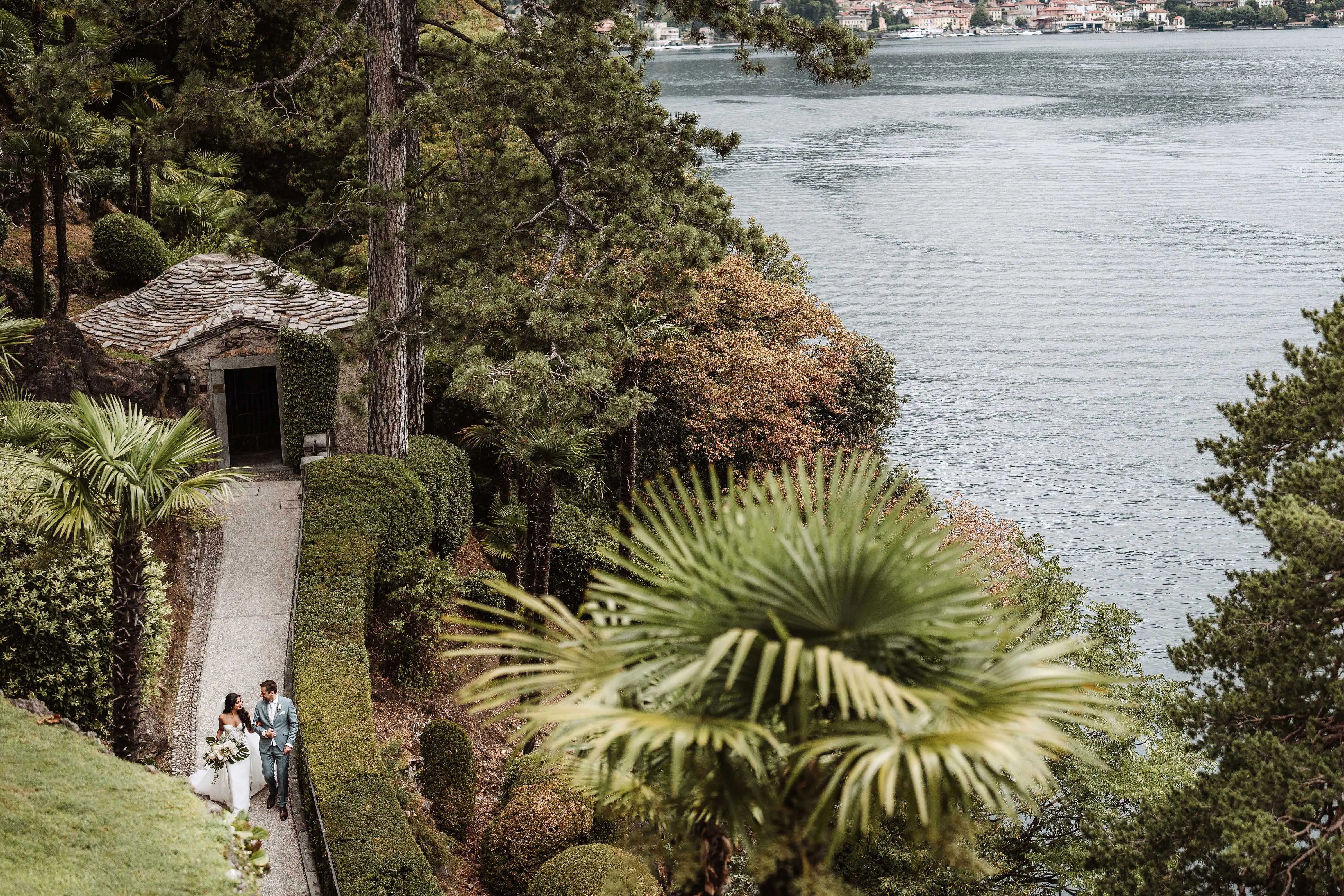 Bride and groom walking hand in hand through a terraced garden overlooking Lake Como during their wedding day