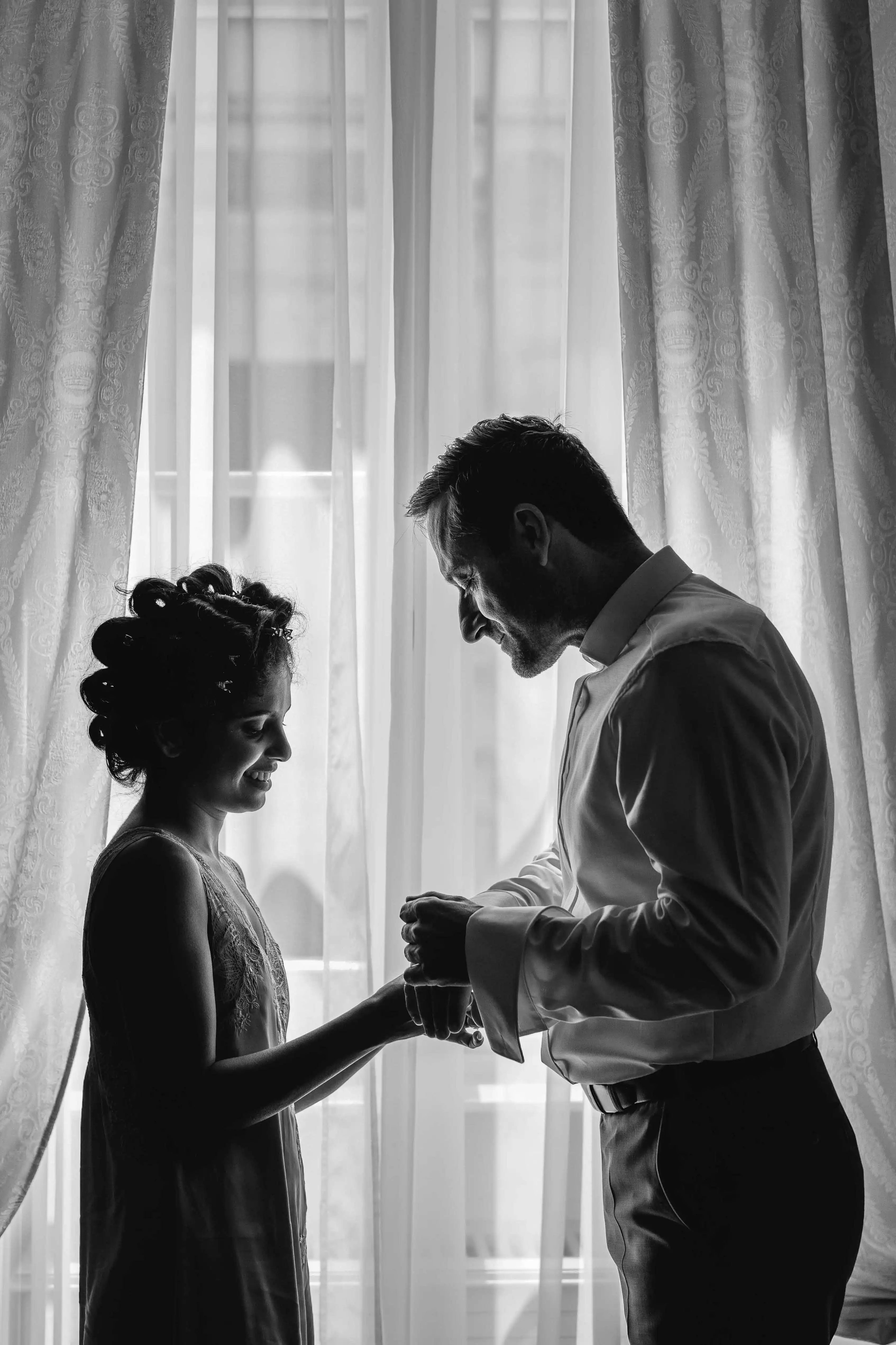 Bride and groom silhouetted by a window holding hands during wedding preparations