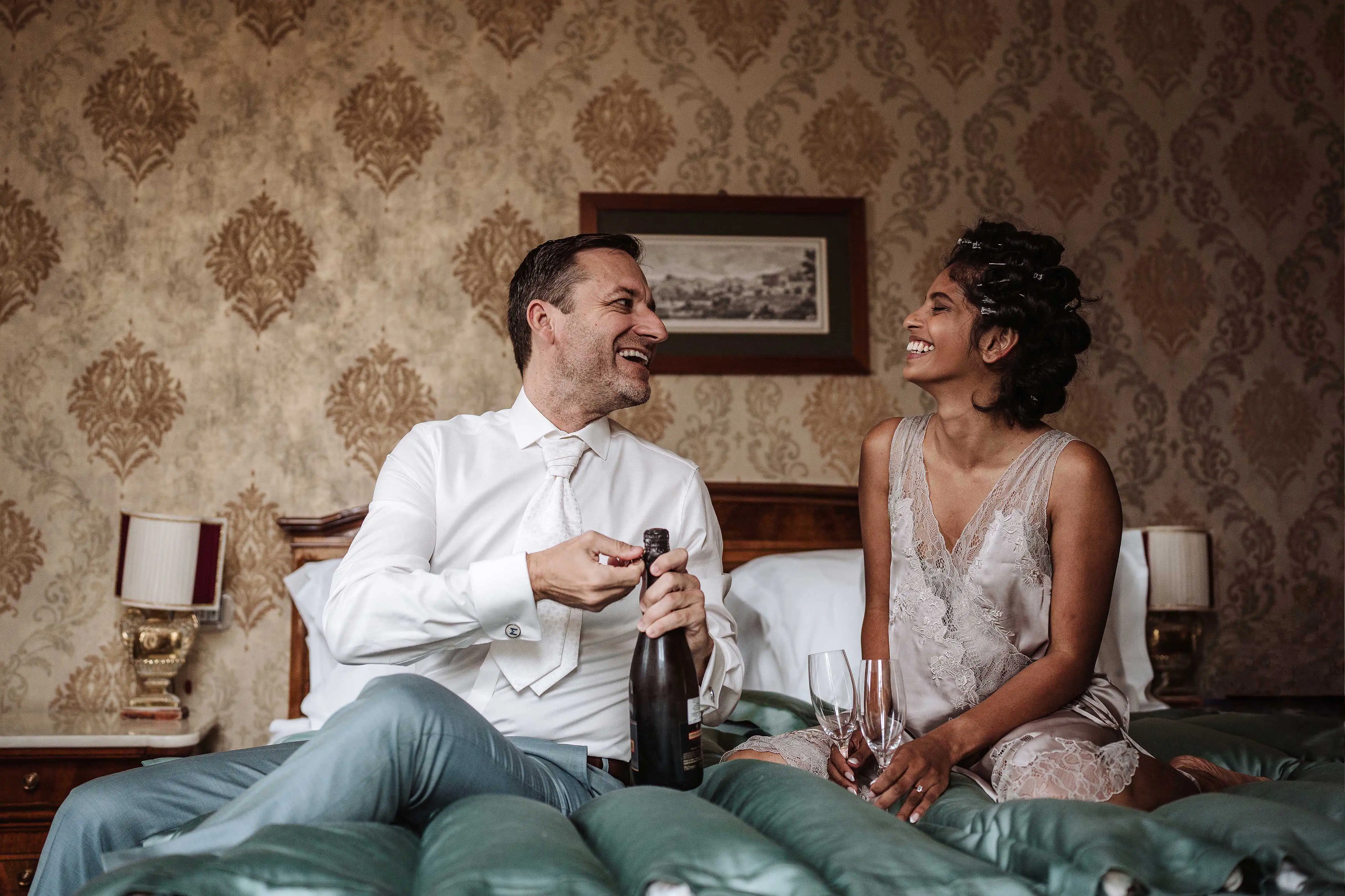 Bride and groom sitting on a bed toasting with champagne during wedding preparations