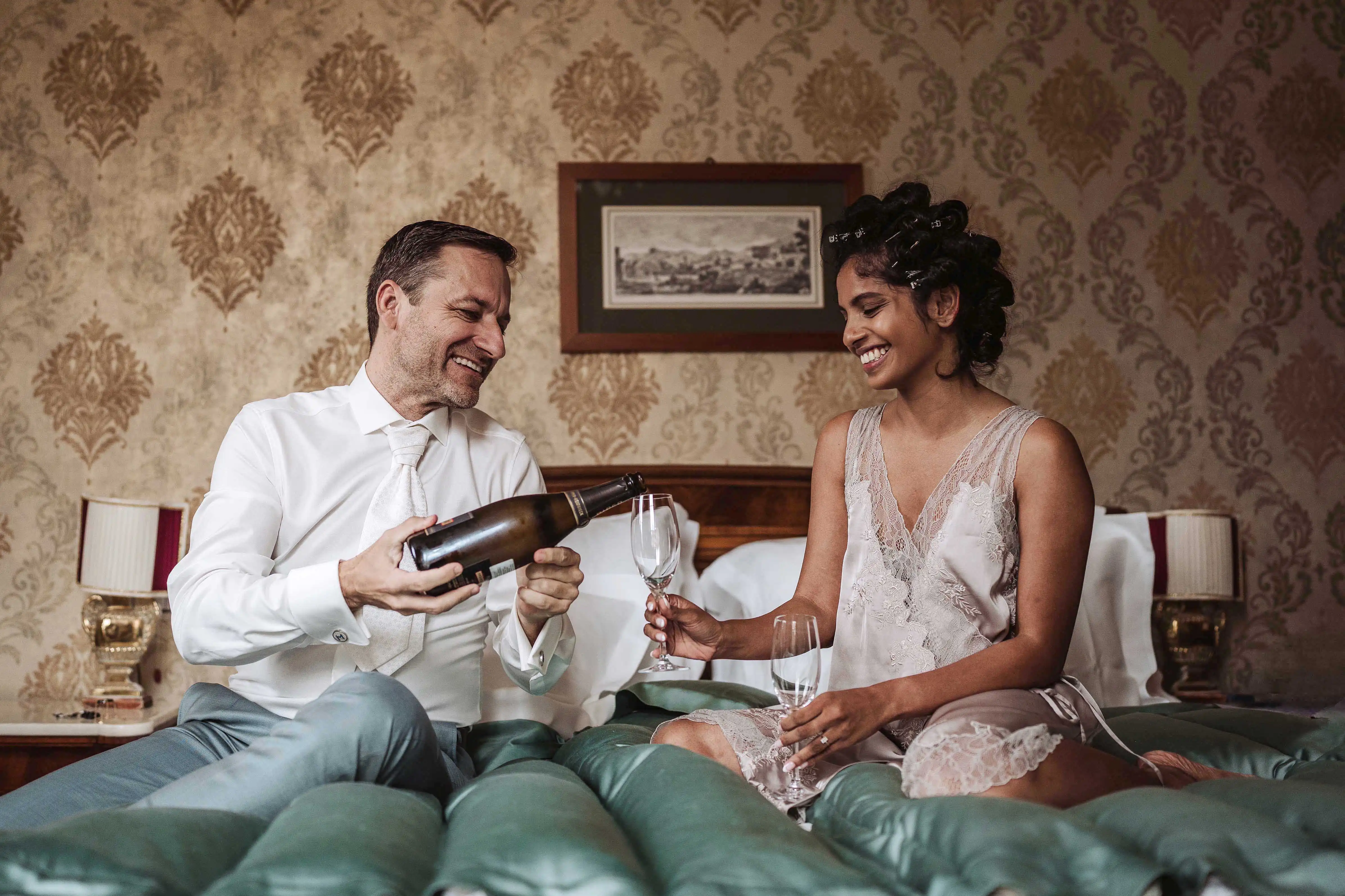 Groom opening a bottle of champagne while bride smiles at him on the bed