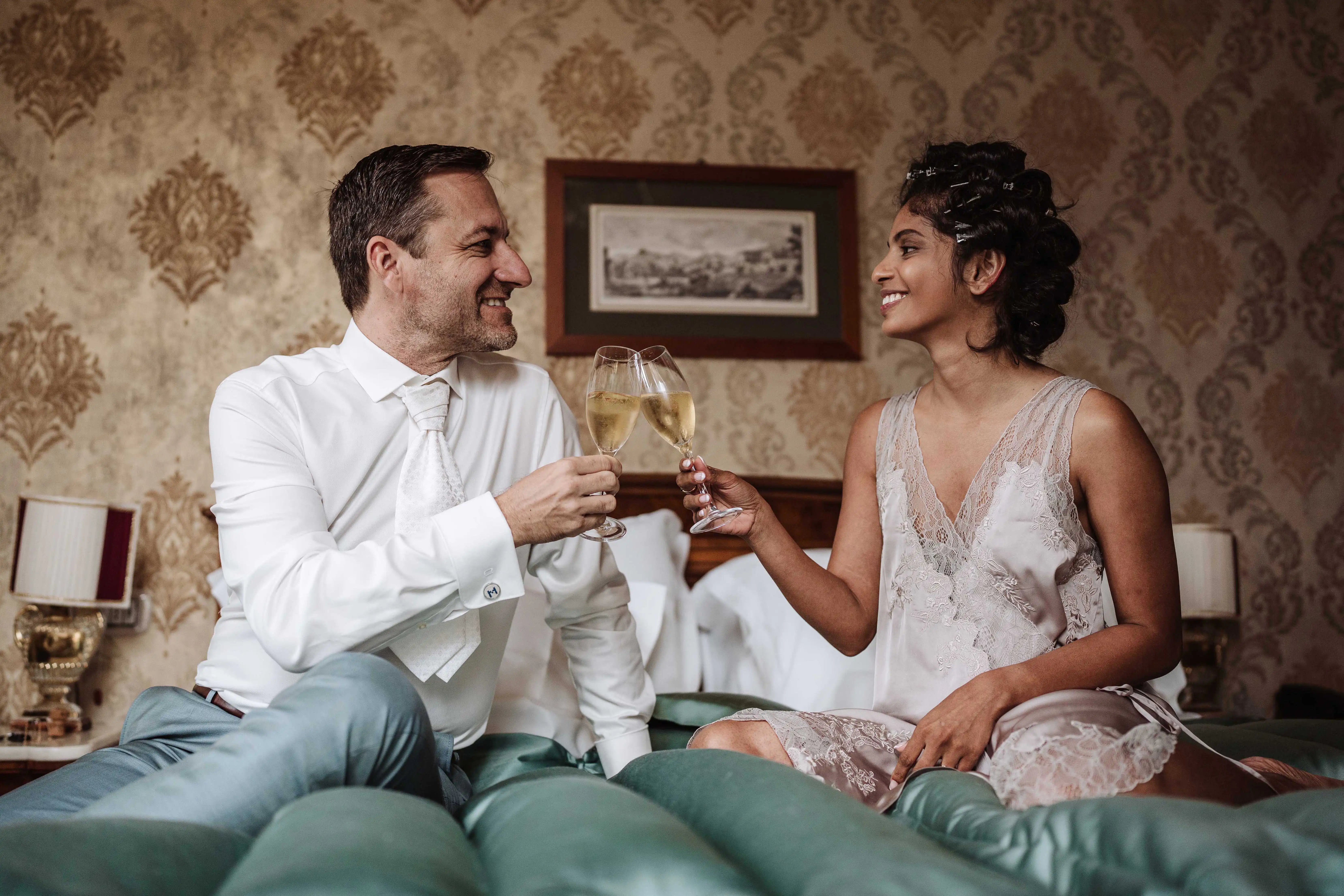 Bride and groom toasting with champagne while sitting on a bed during wedding preparations