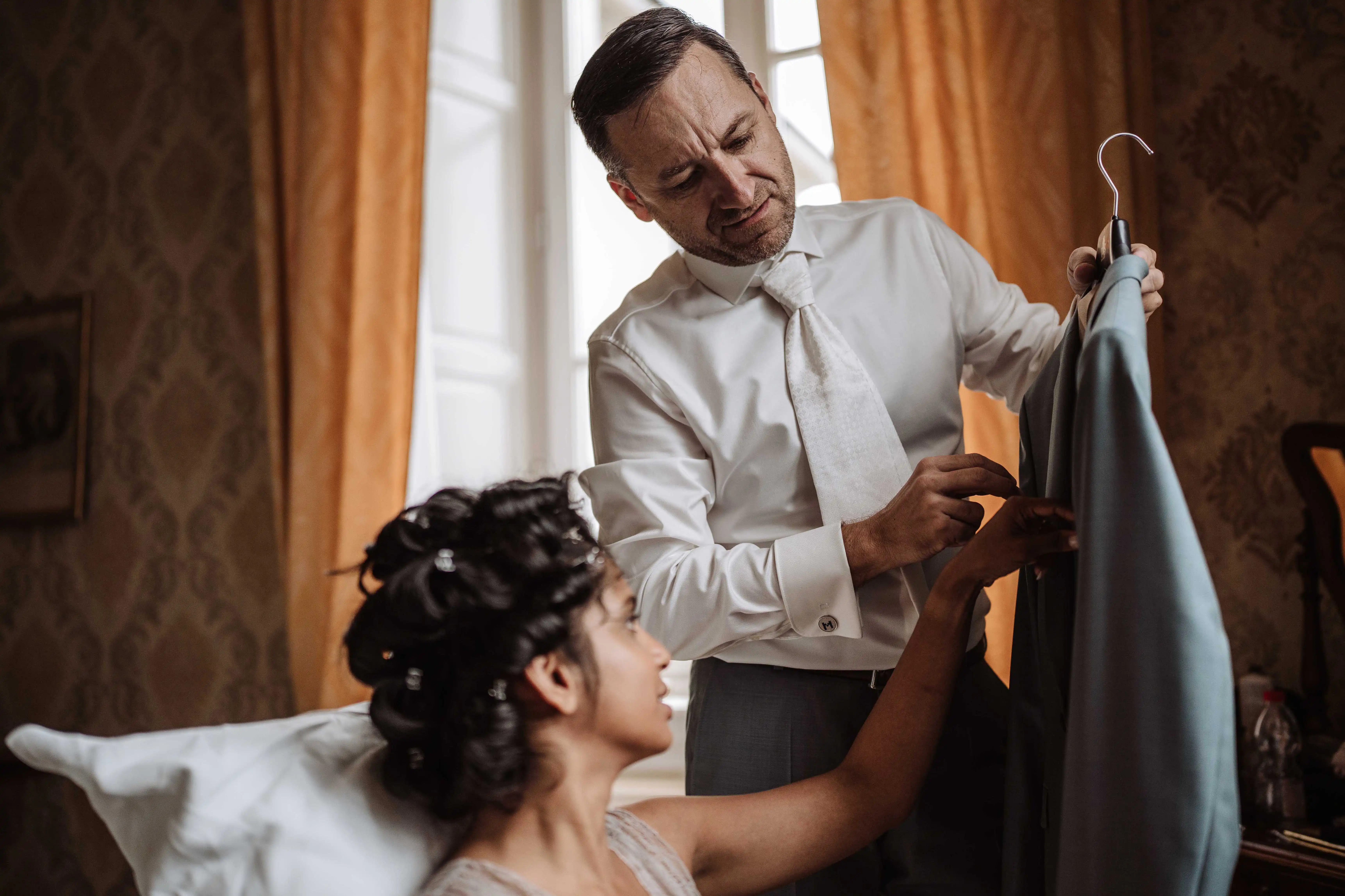 Groom helping bride with dress while holding suit jacket in elegant room