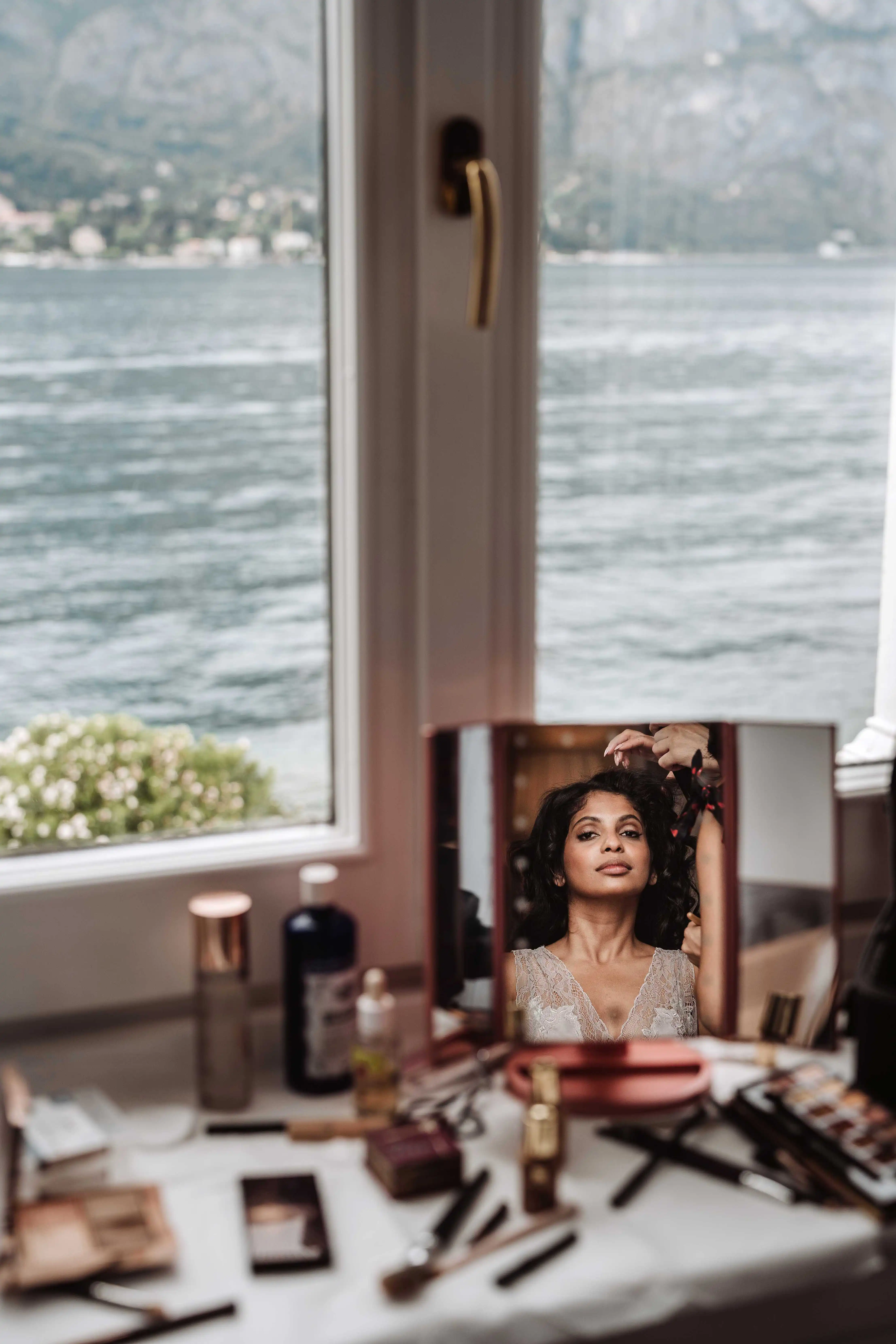 Bride reflected in mirror while hairstylist works near window with lake view