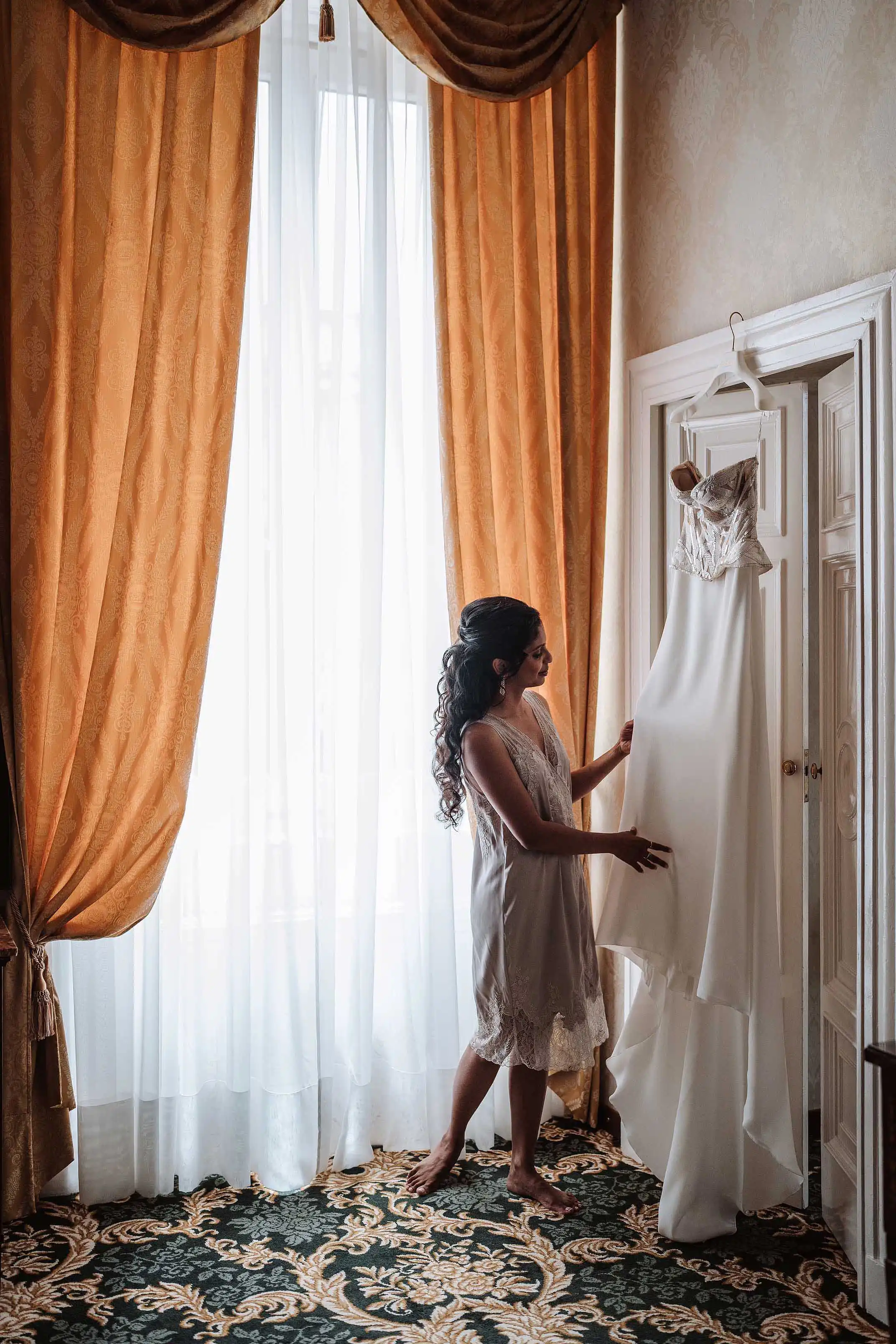 Bride standing by window looking at her wedding dress hanging on wardrobe
