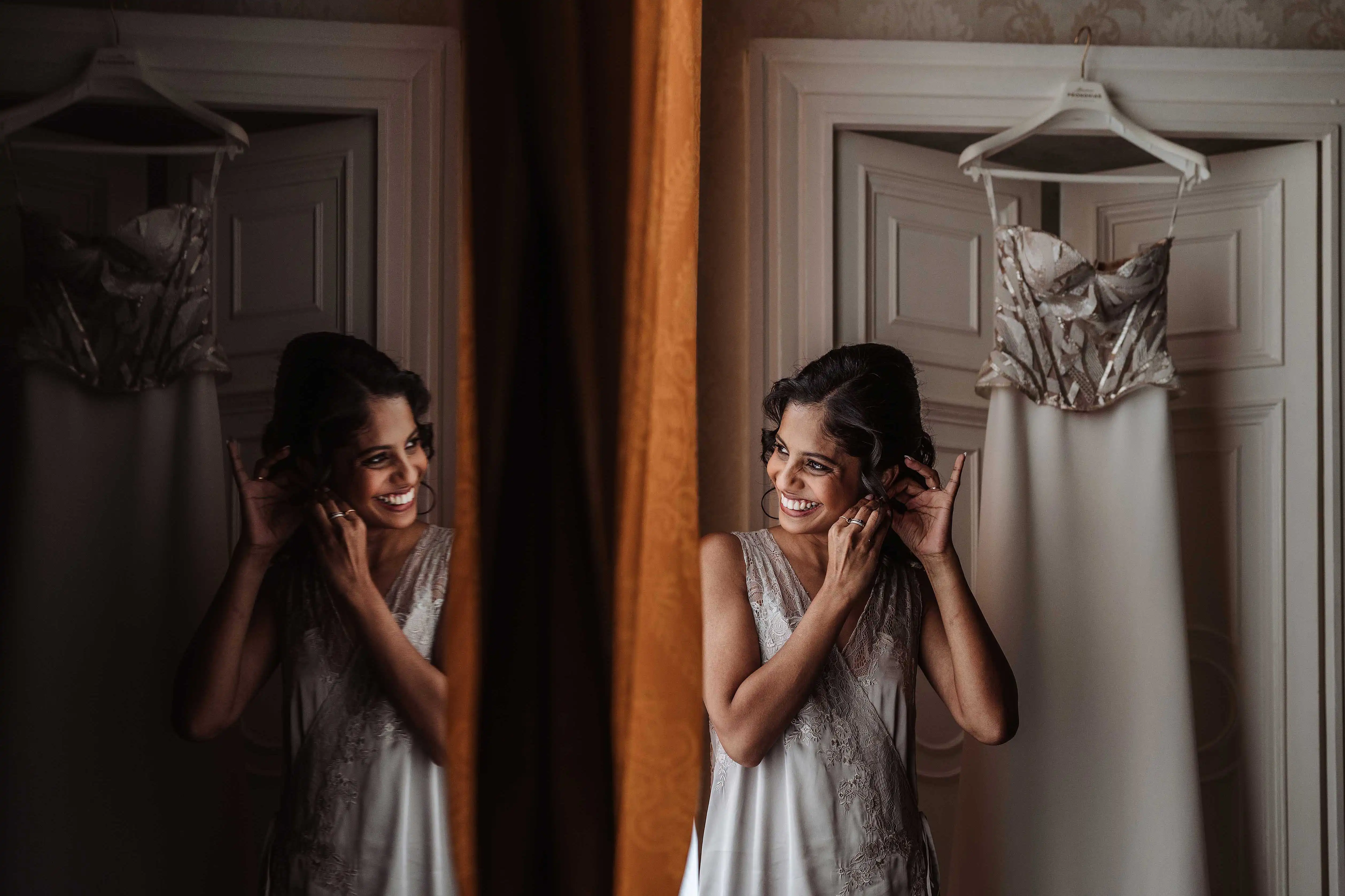Bride smiling while putting on earrings reflected in mirror with dress behind