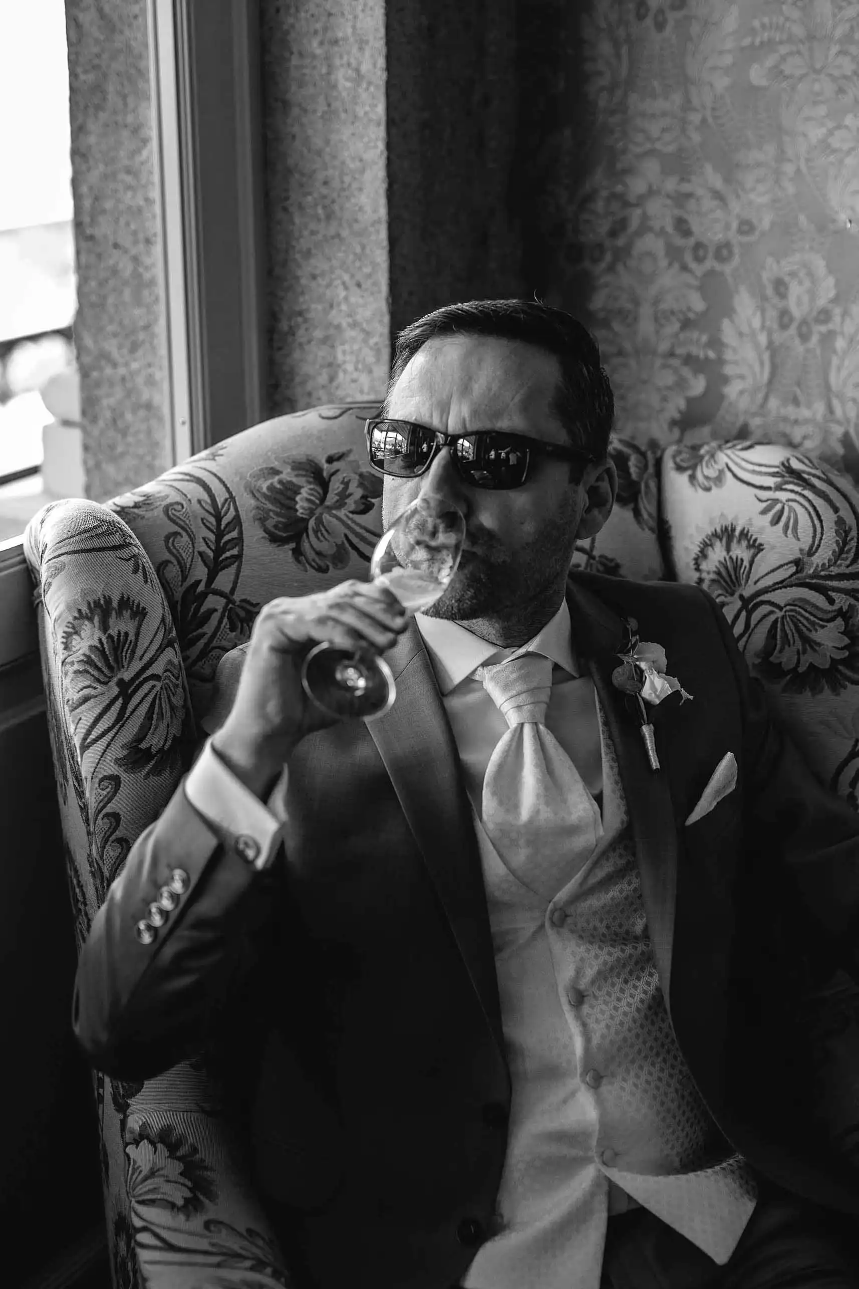 Groom in suit and sunglasses relaxing with a drink in vintage armchair before wedding