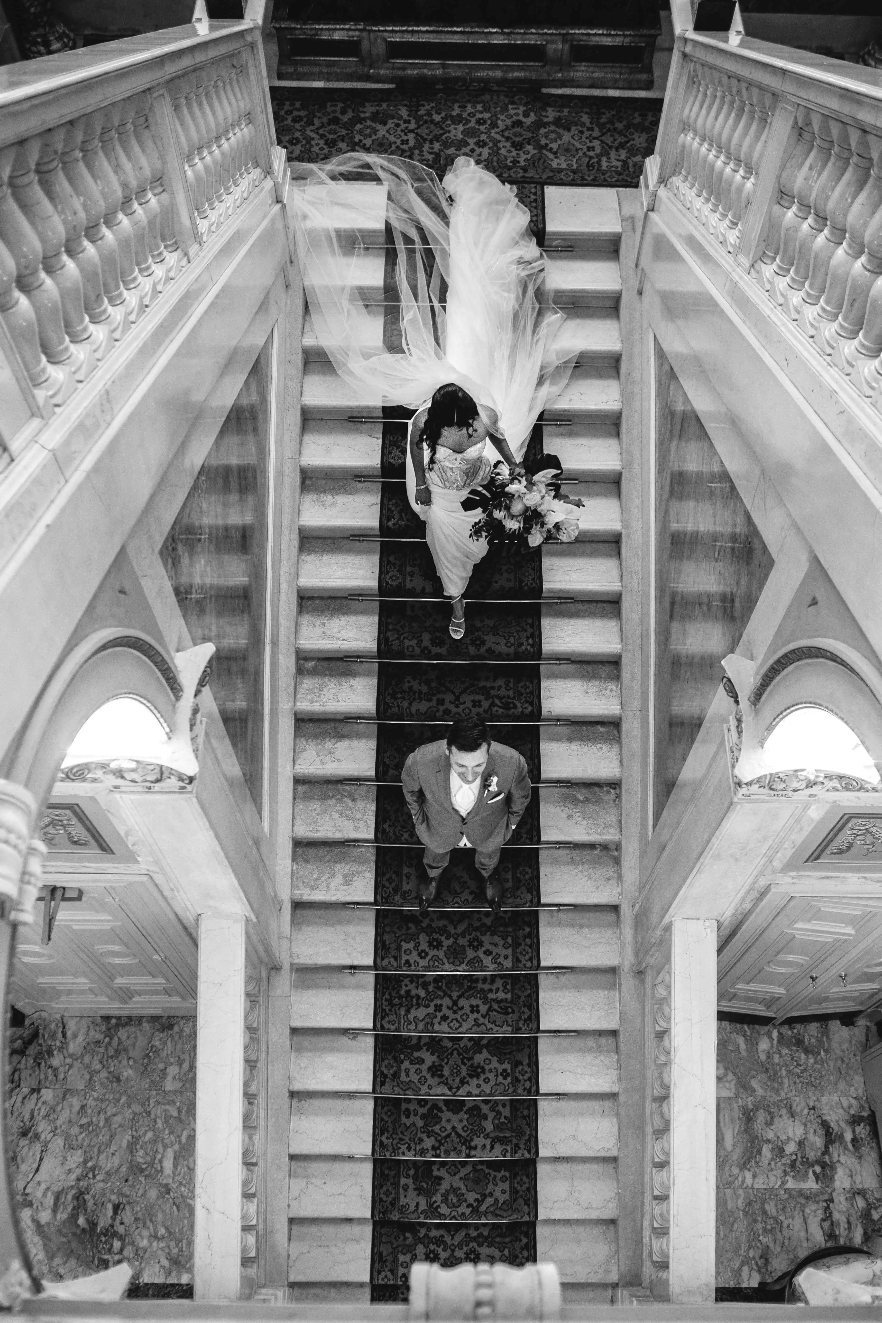 Bride walking down grand staircase toward groom in elegant black and white scene