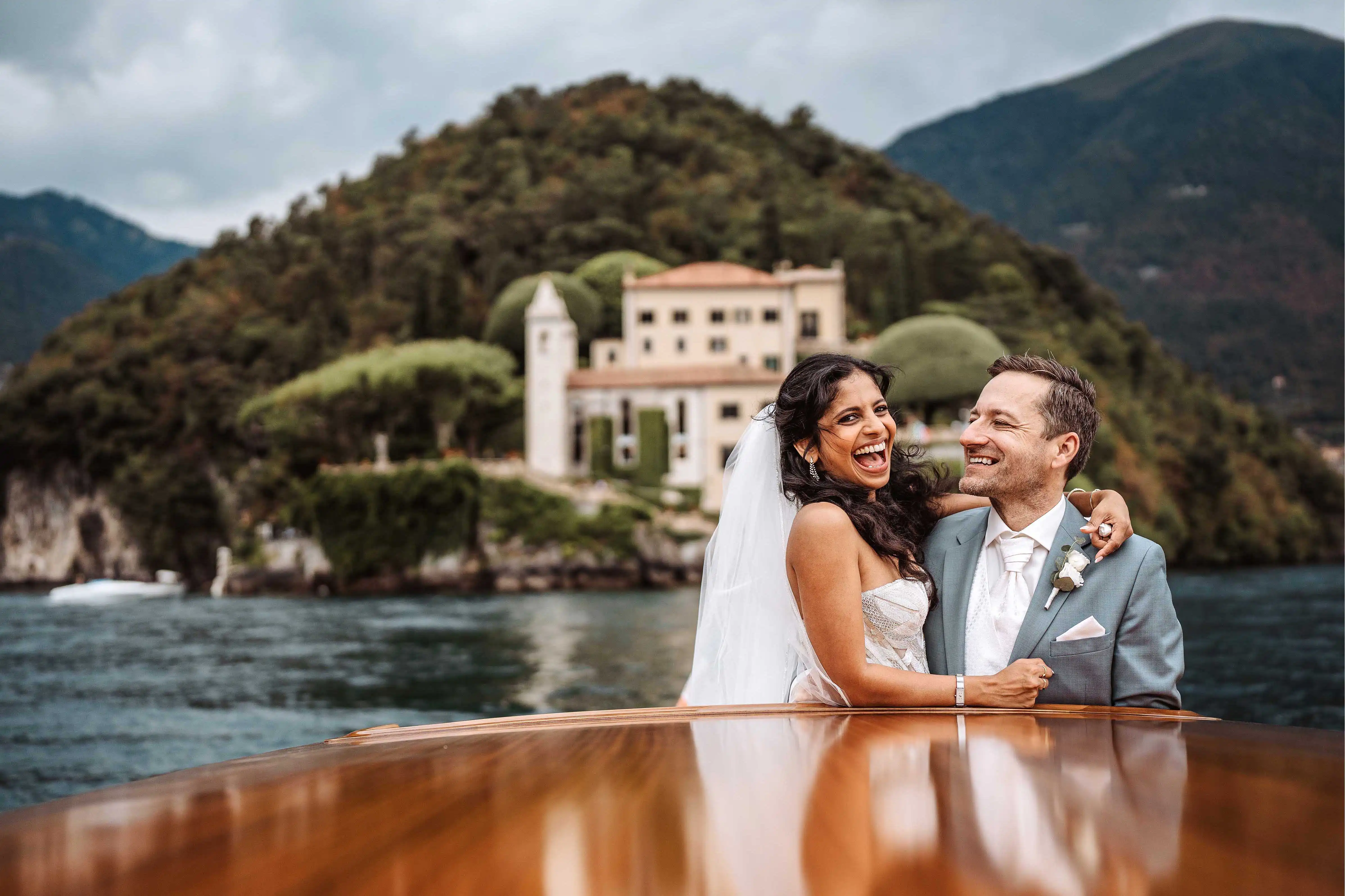 Happy bride and groom laughing on wooden boat on Lake Como with scenic villa behind