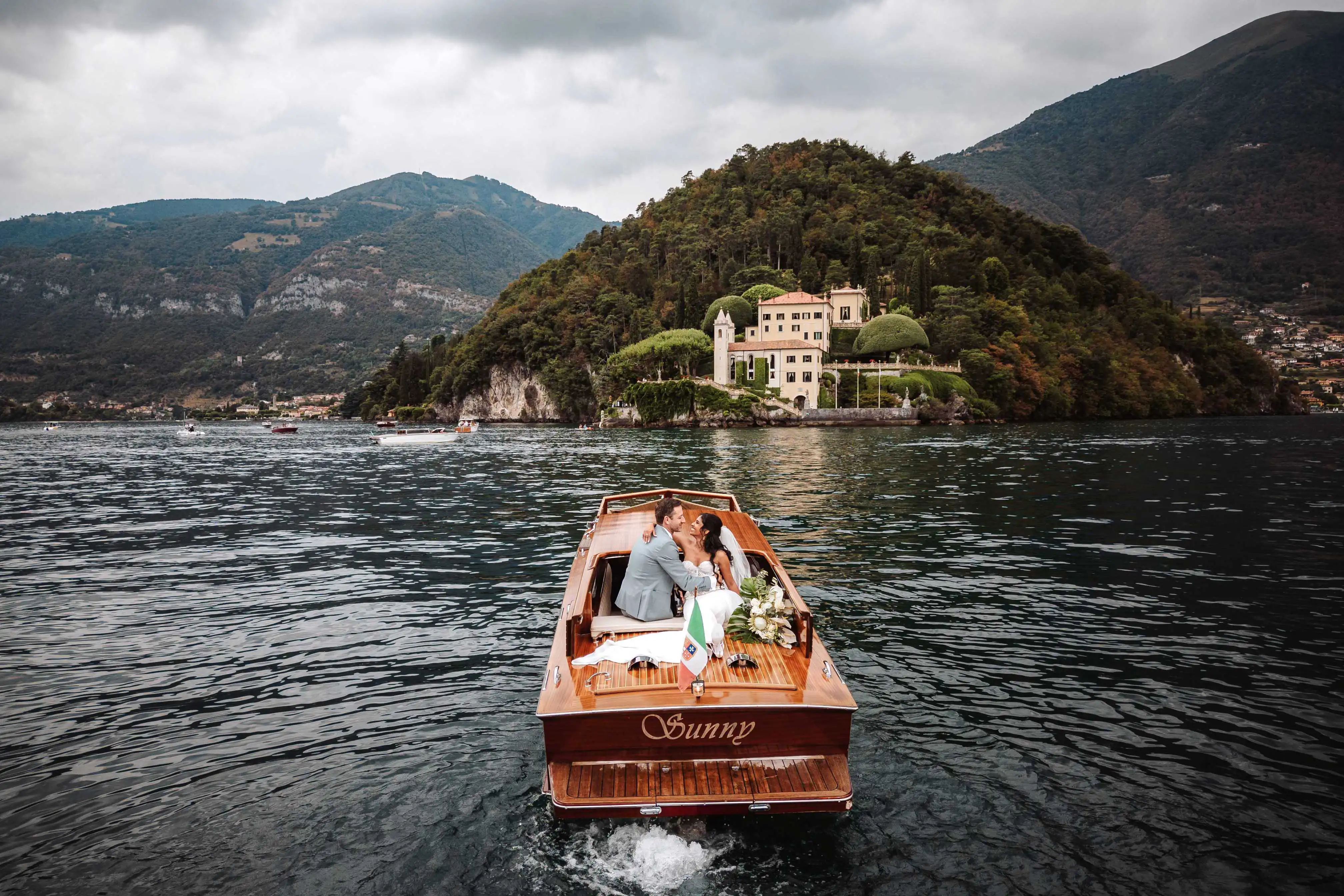 Bride and groom embracing on a wooden boat on Lake Como with Villa del Balbianello in the background under a cloudy sky