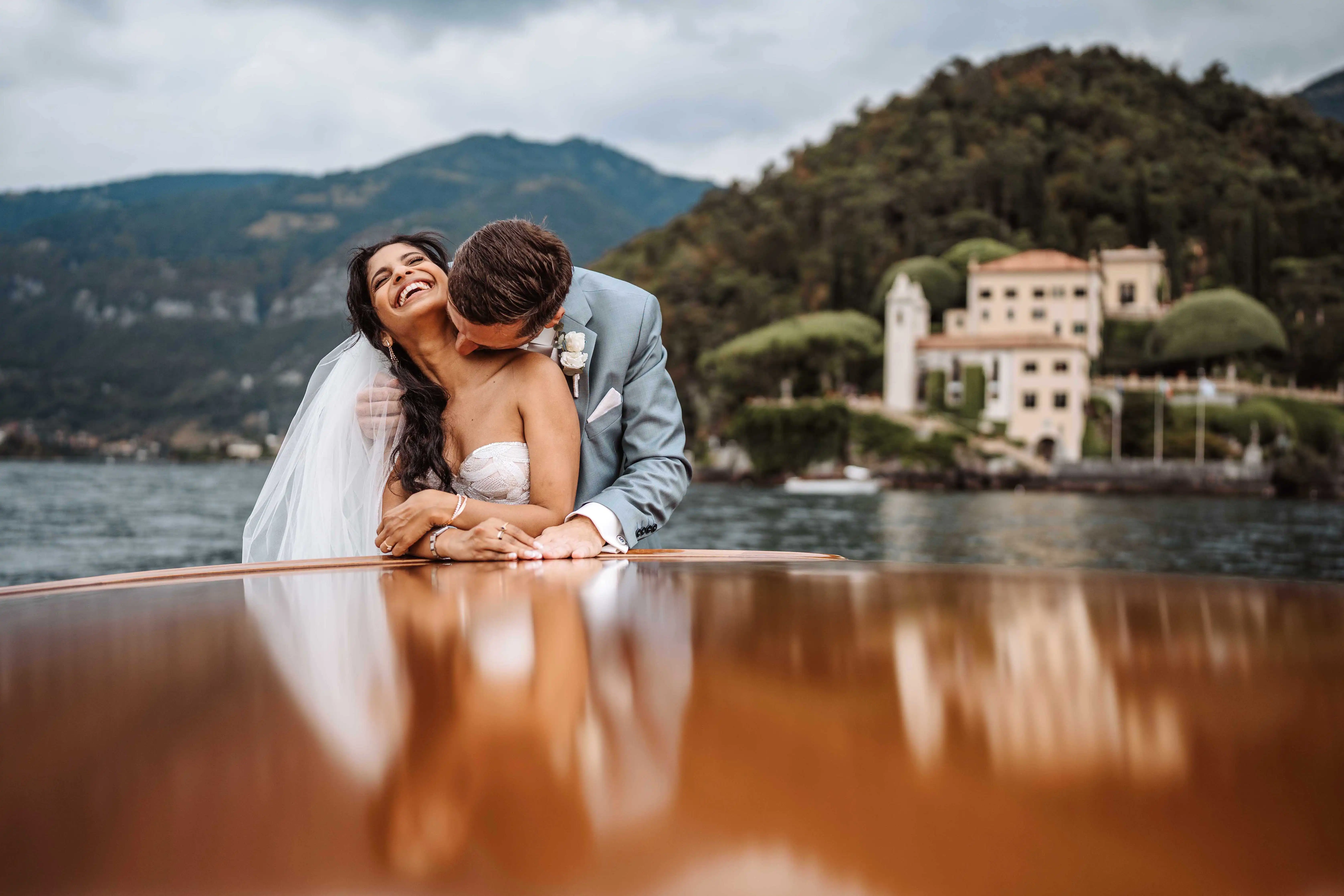 Close-up of joyful bride laughing as groom kisses her on a boat with Lake Como and villa softly blurred behind