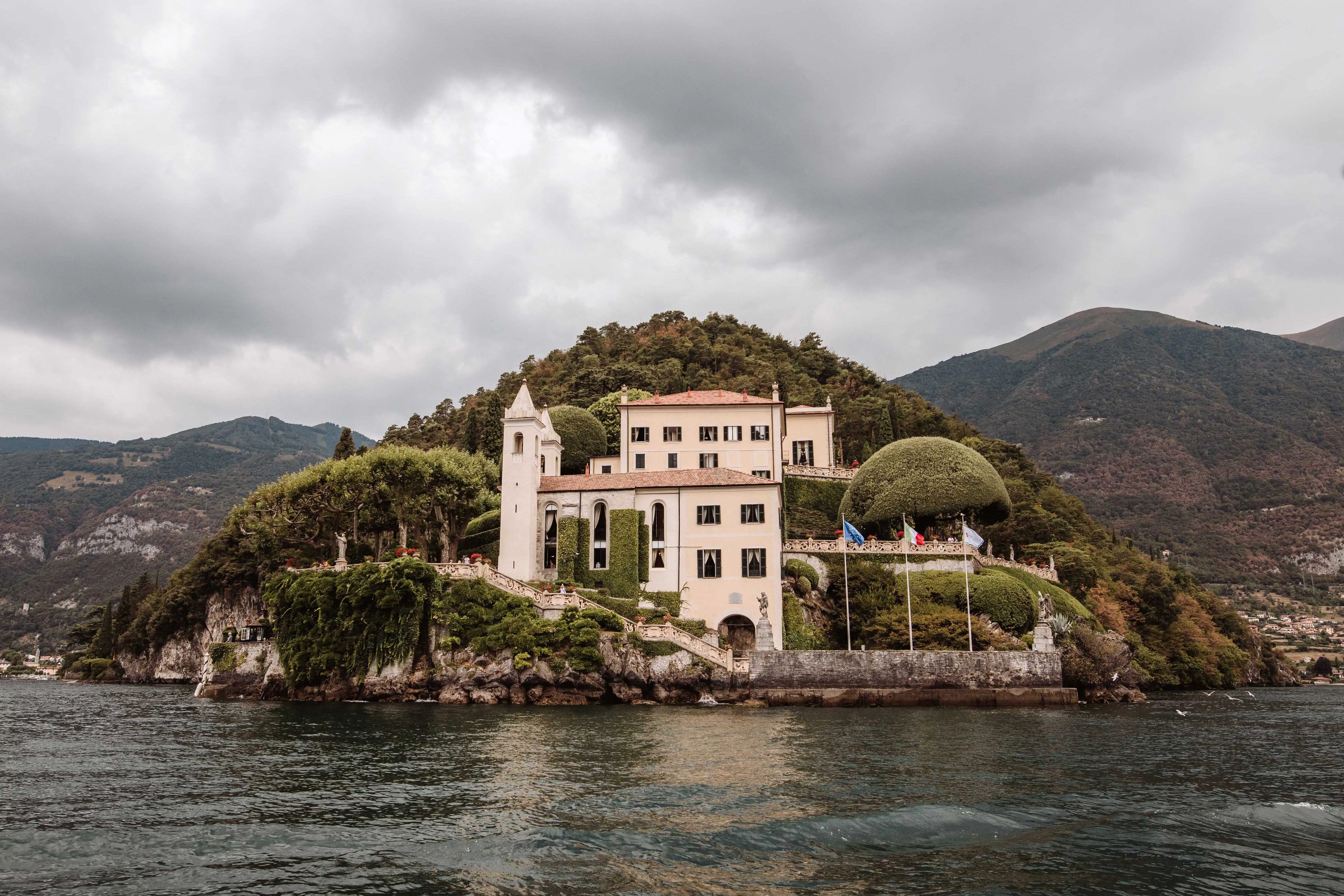 Scenic view of Villa del Balbianello surrounded by lush greenery on Lake Como under dramatic clouds
