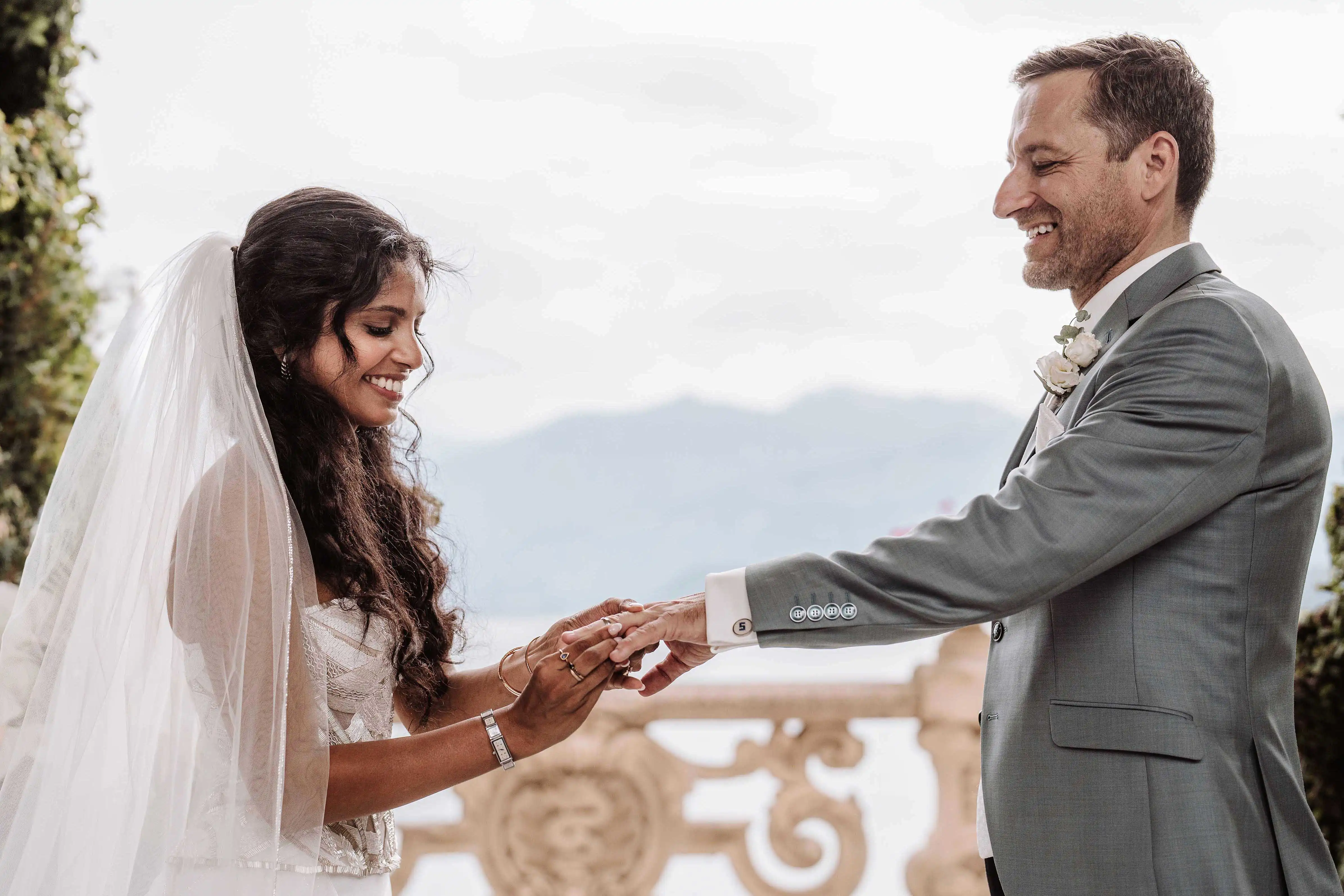 Bride and groom smiling during ring exchange with soft lake view in the background