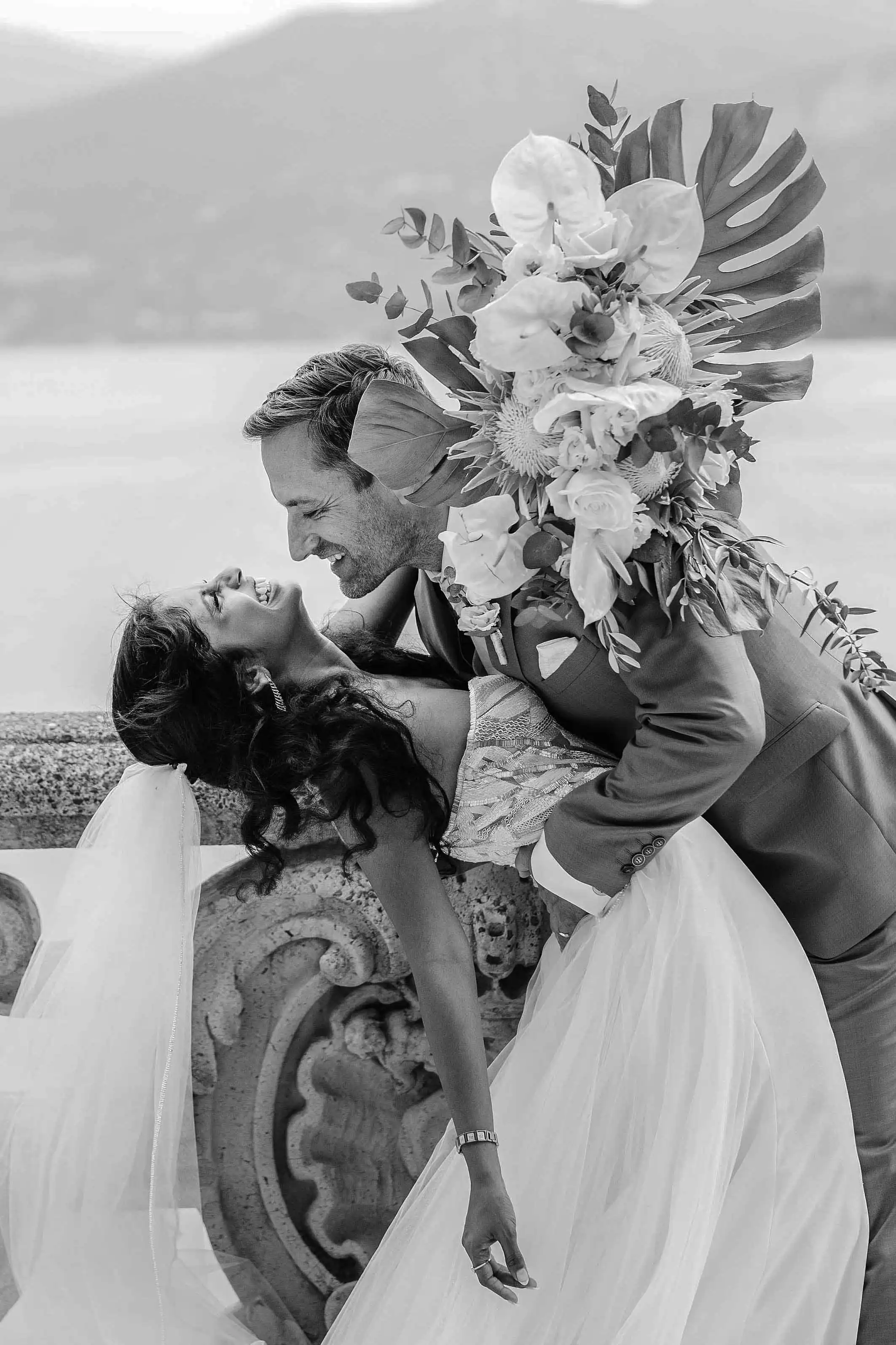 Groom holding the bride and smiling with a large bouquet in a romantic black and white portrait by Lake Como