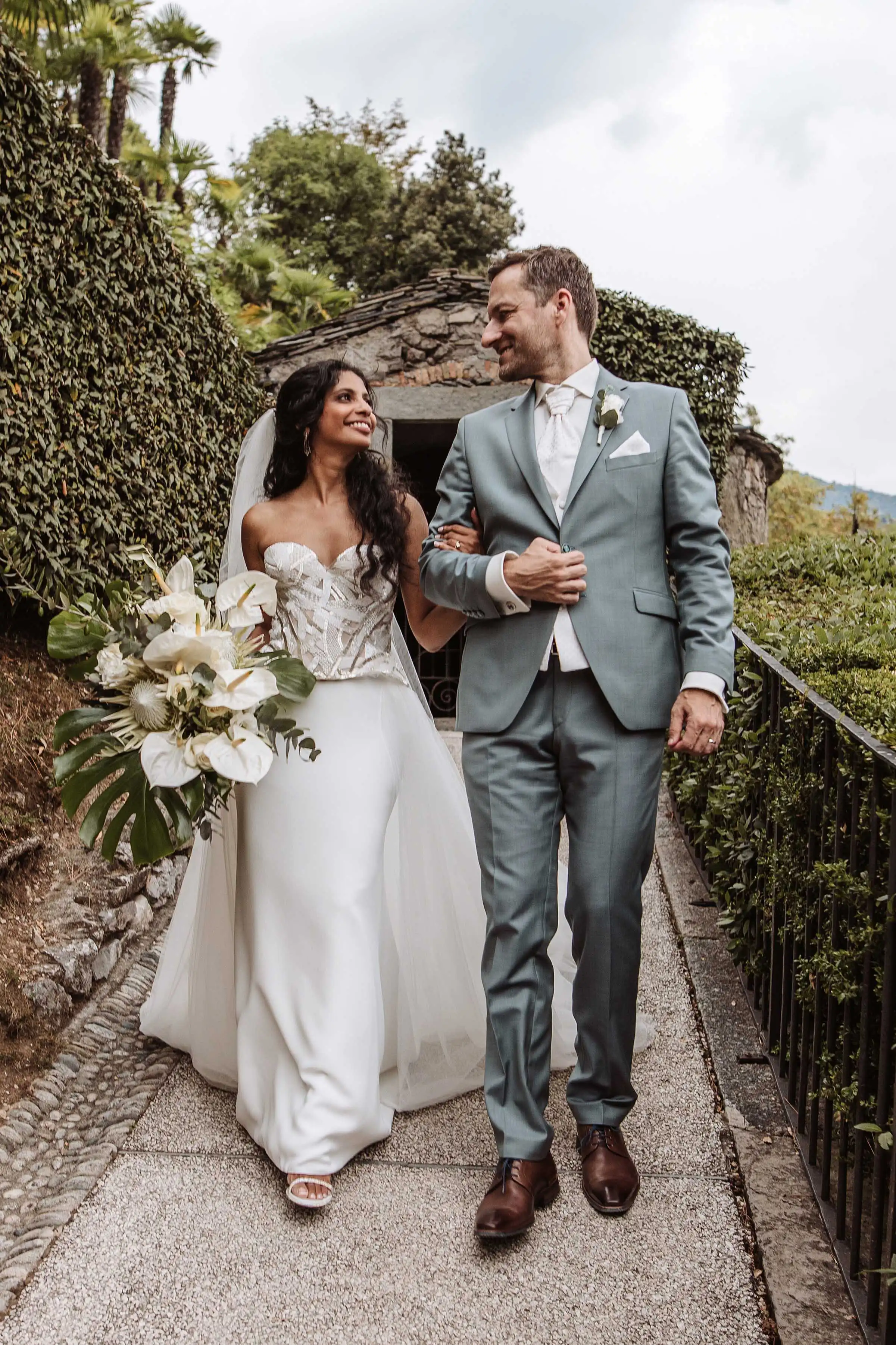 Elegant couple walking along a garden path with tropical bouquet at a luxury Lake Como wedding venue