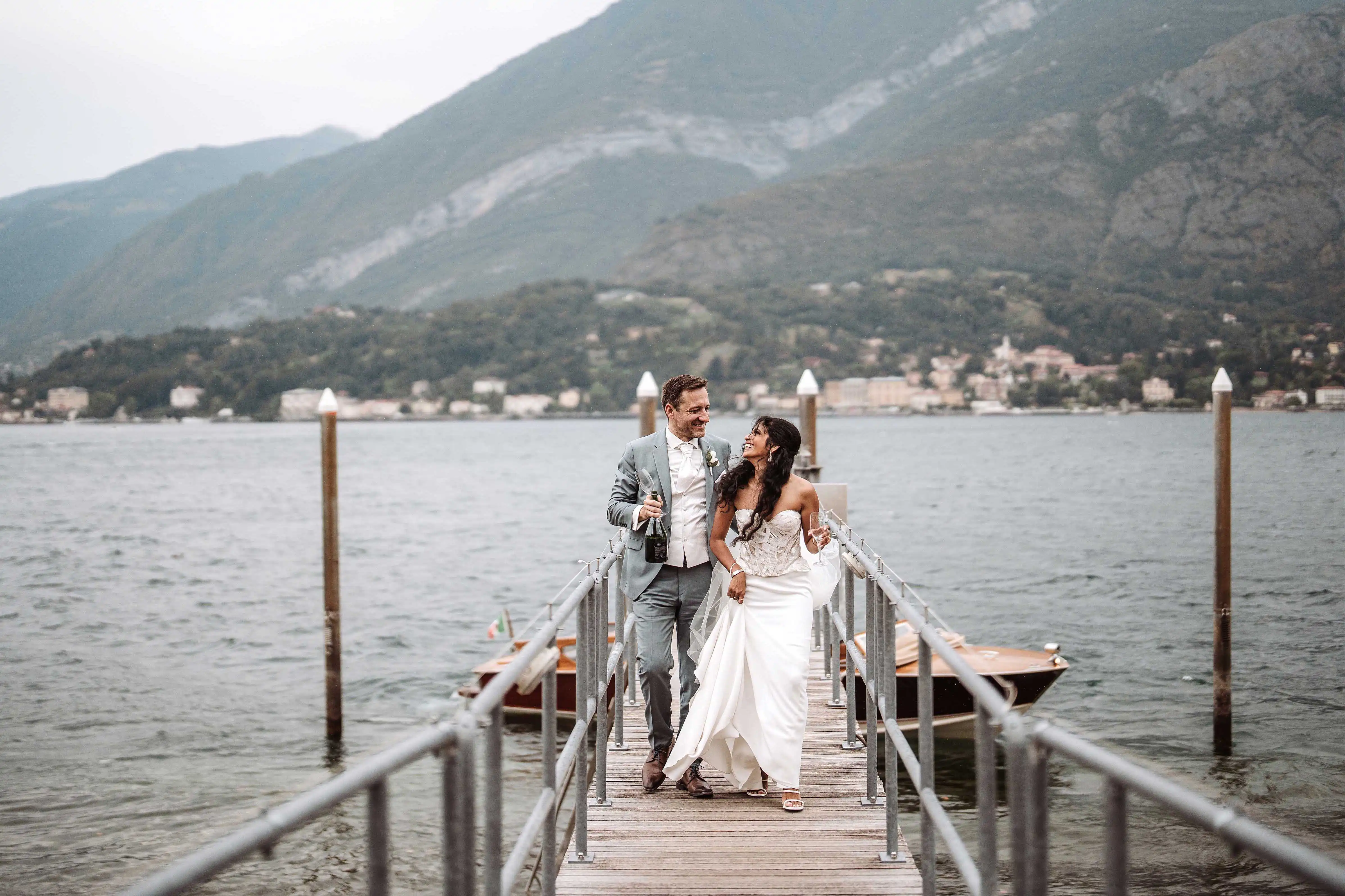 Bride and groom walking on a wooden pier by Lake Como holding champagne and smiling at each other during an intimate wedding moment