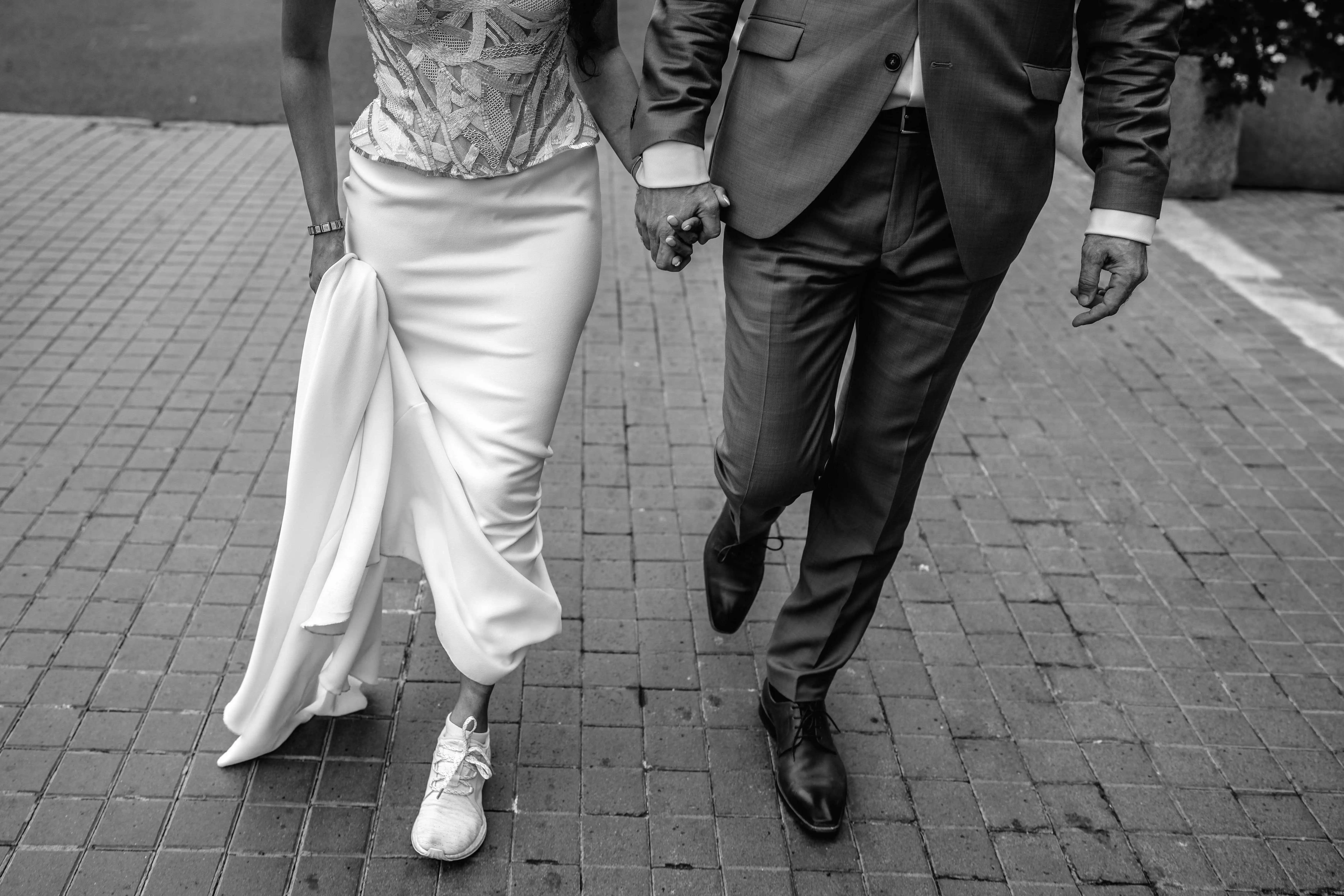 Close-up black and white photo of bride and groom holding hands while walking, highlighting elegant wedding dress and suit details