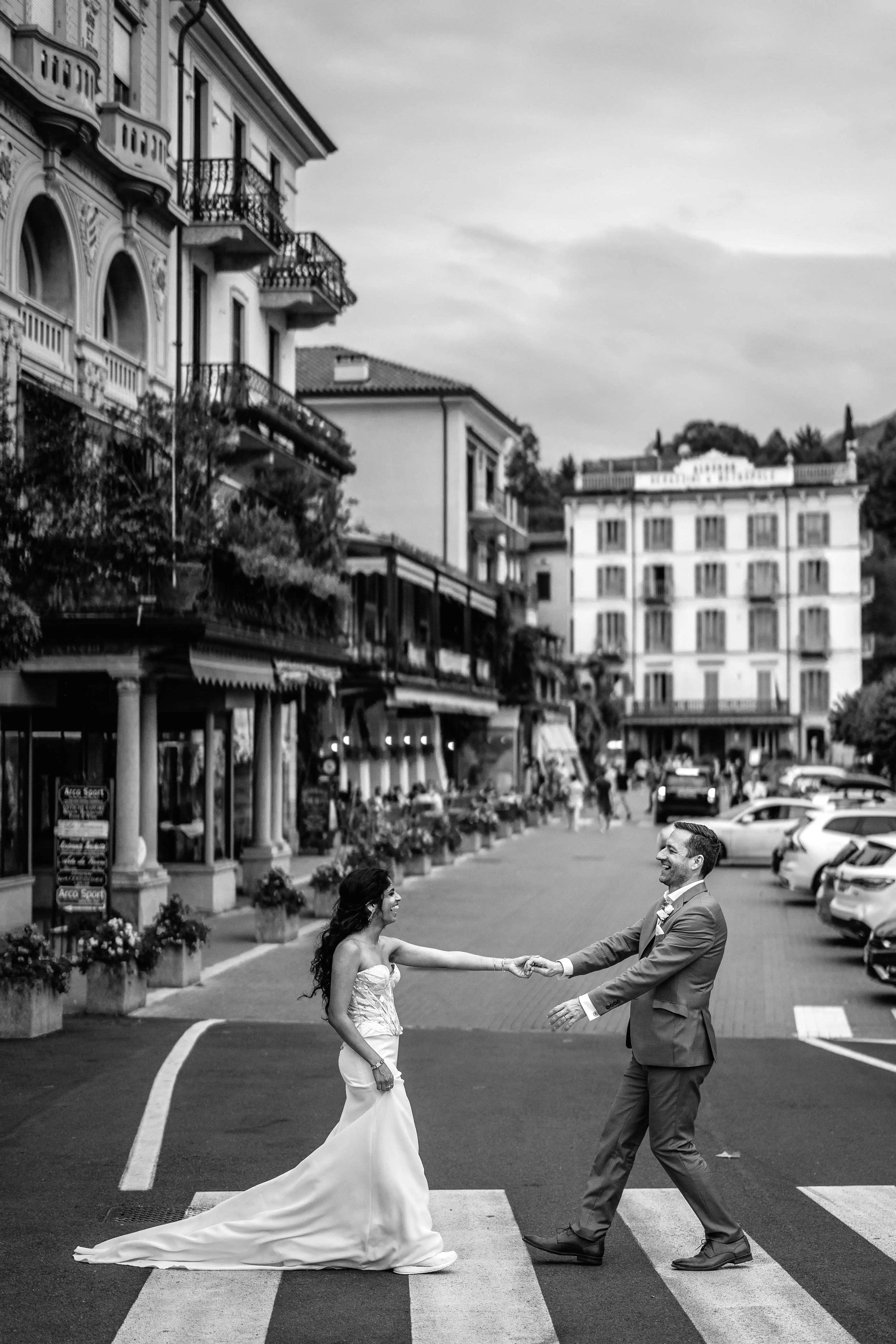 Bride and groom playfully holding hands across a crosswalk in a Lake Como town with historic Italian architecture in the background