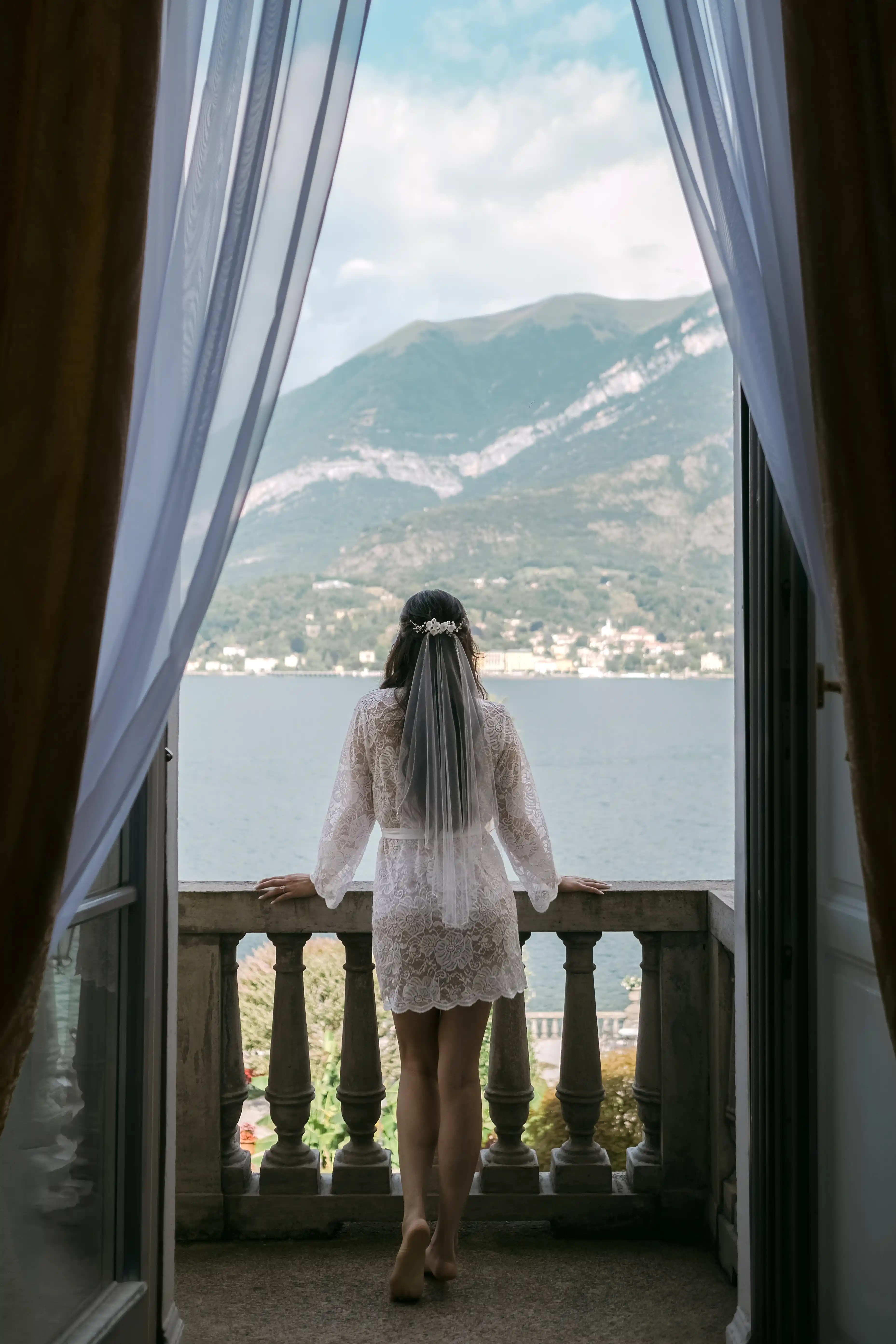 Bride in lace robe looking out over Lake Como from a historic villa balcony, intimate pre-ceremony moment