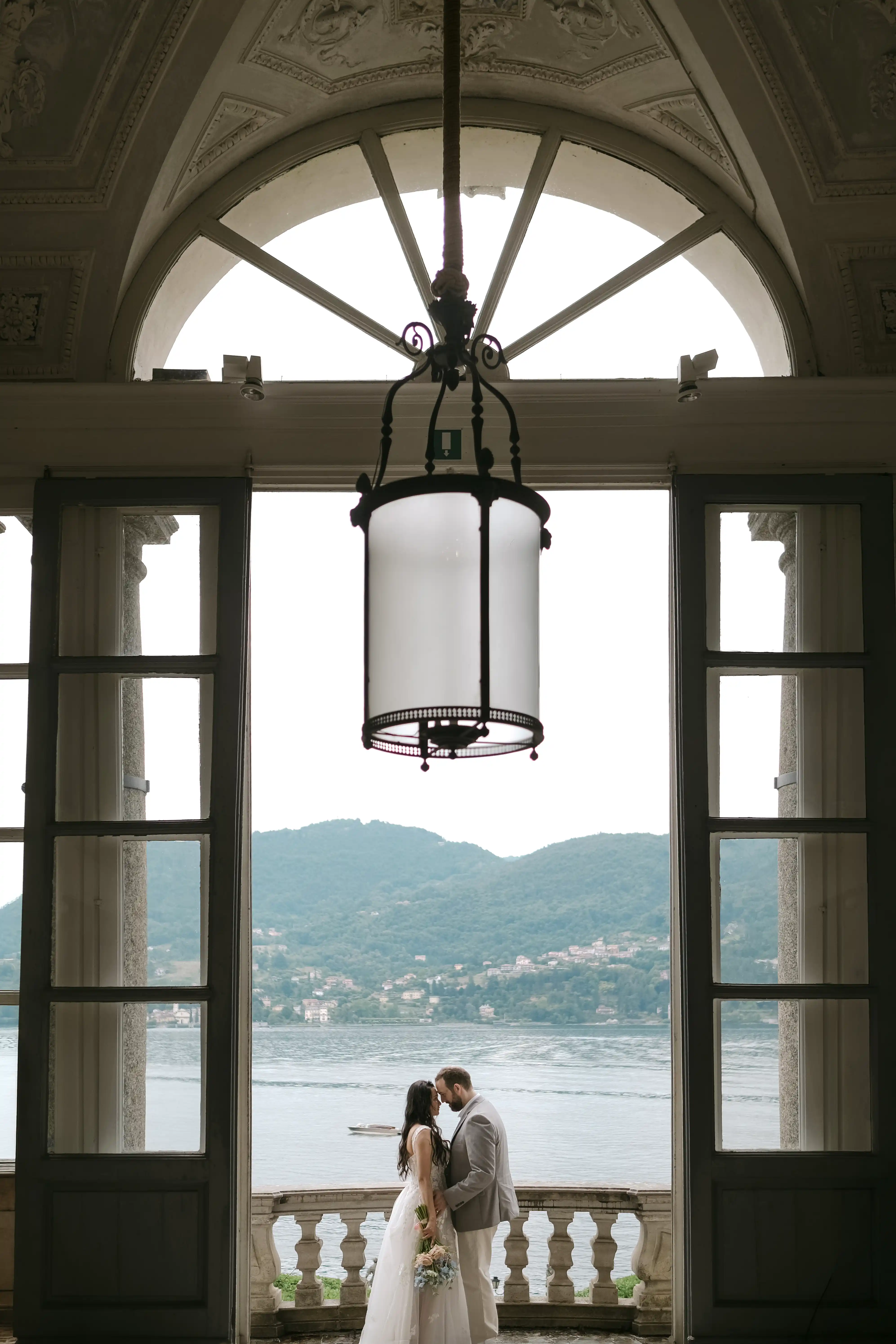 Bride and groom embracing on a terrace overlooking the lake, framed by elegant architecture and soft natural light