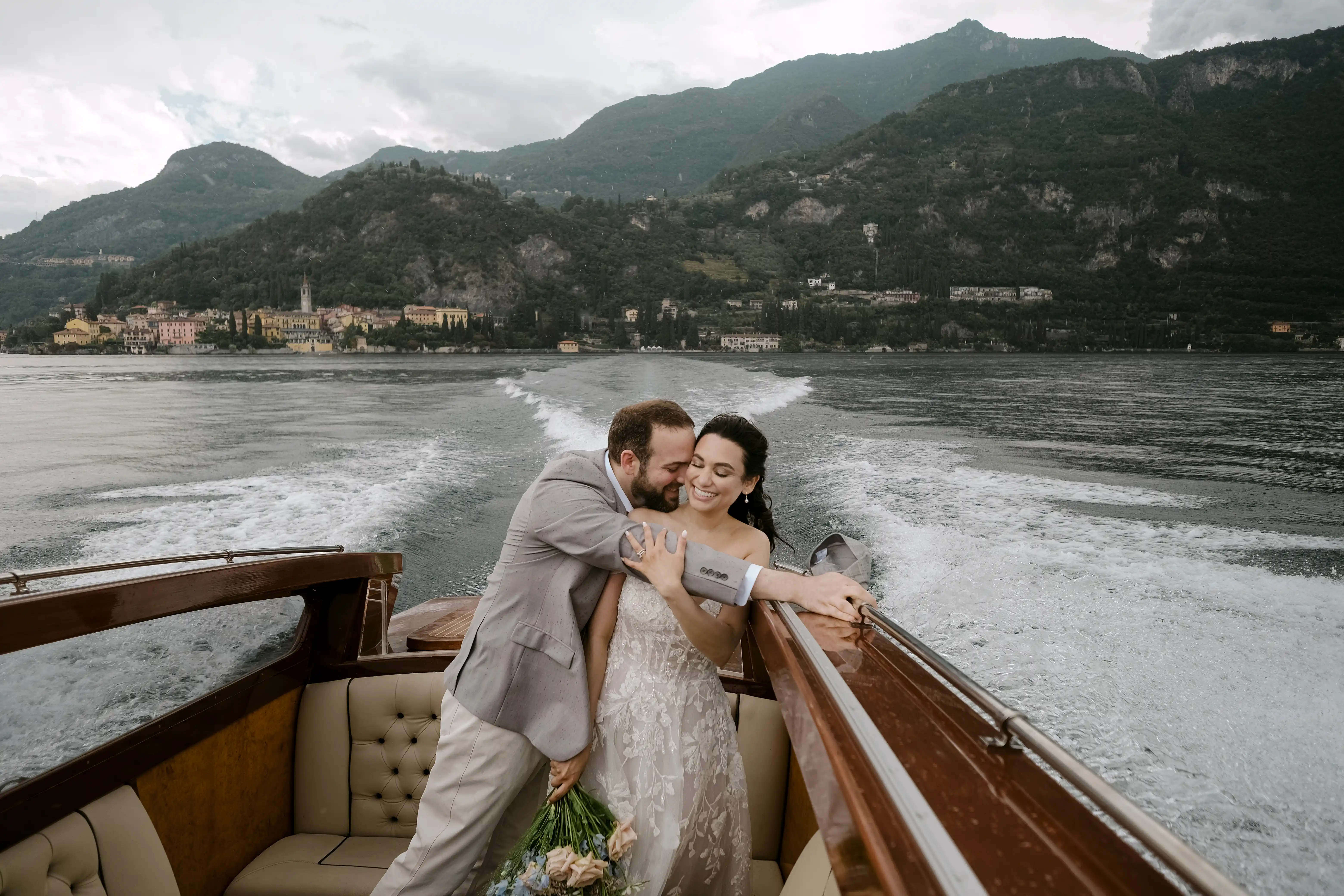 Bride and groom embracing on a wooden boat speeding across Lake Como, joyful and intimate moment with mountains in the background