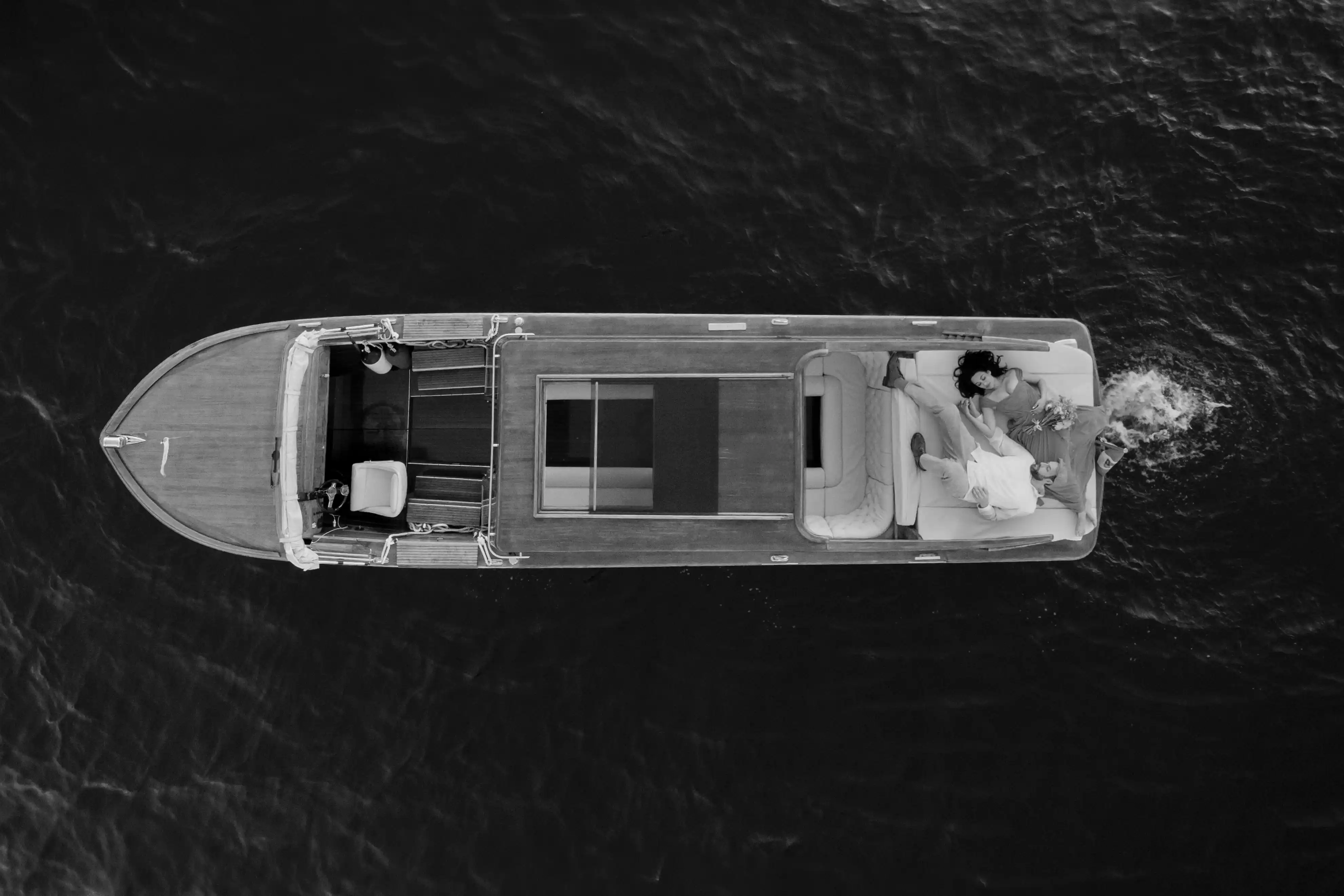 Aerial black and white view of a couple relaxing on a classic wooden boat in the middle of Lake Como, cinematic and intimate elopement scene