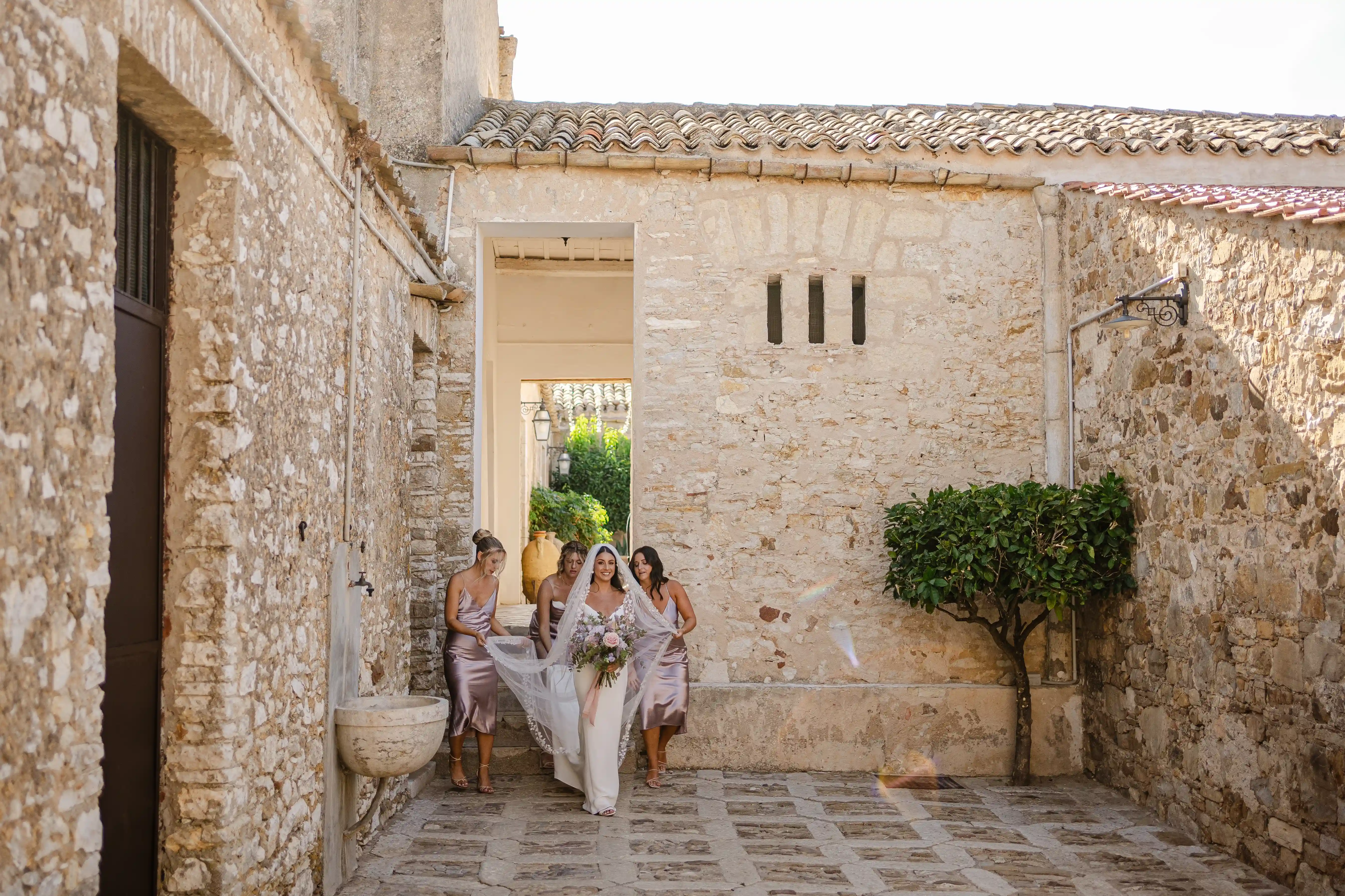 Bride walking through a rustic Sicilian courtyard with bridesmaids adjusting her veil
