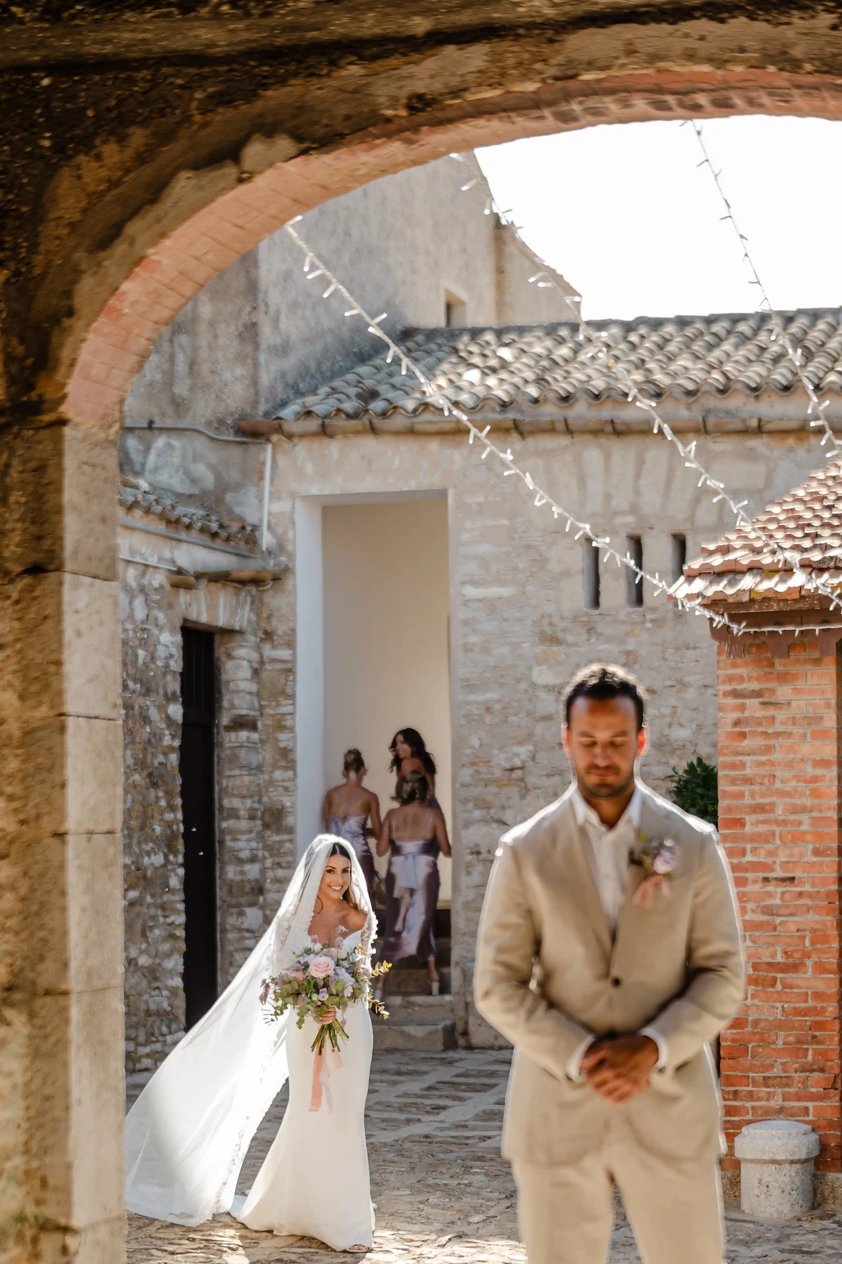 Groom waiting during first look as bride approaches through stone archway in Sicily