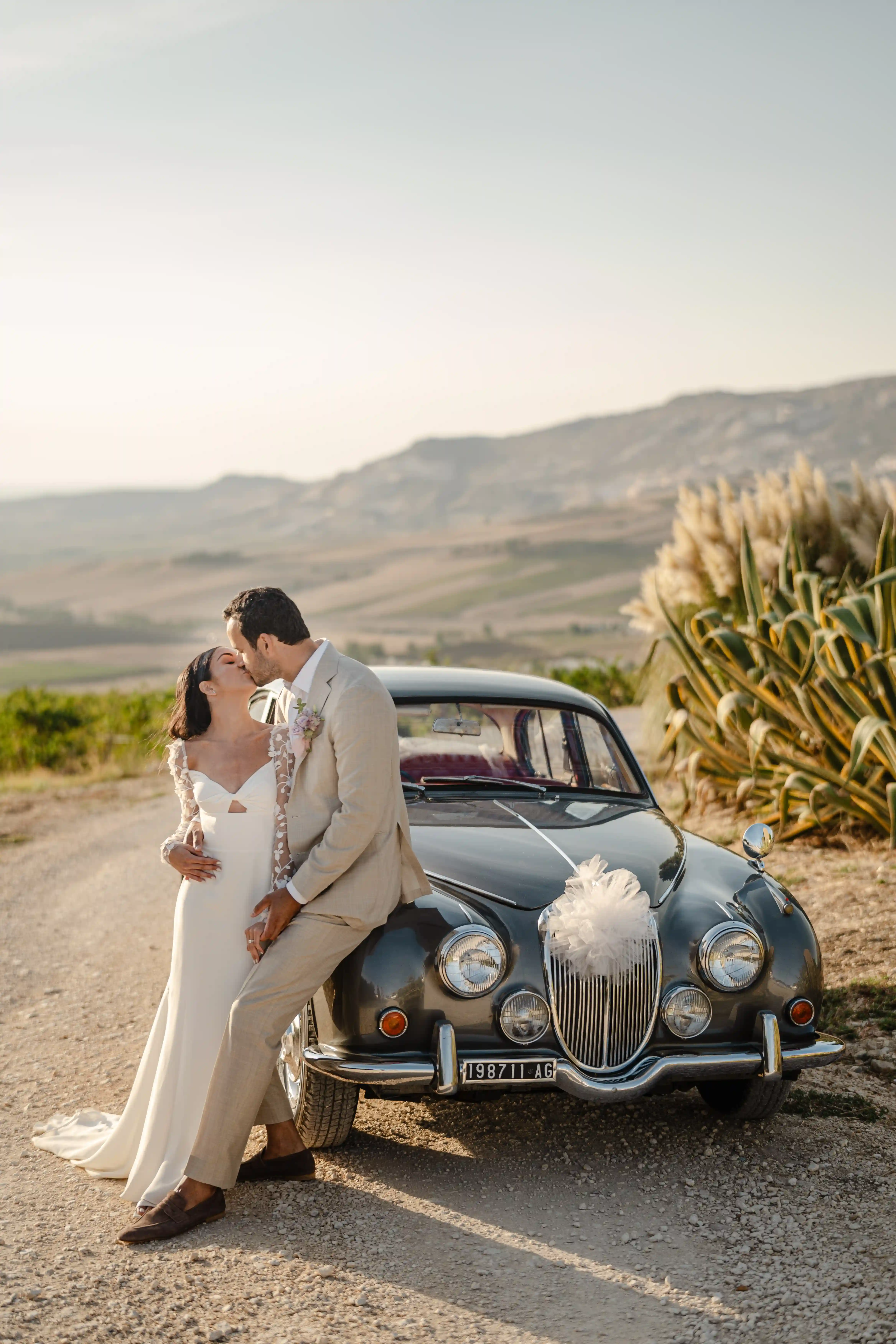 Bride and groom kissing beside vintage car in Sicilian countryside at sunset