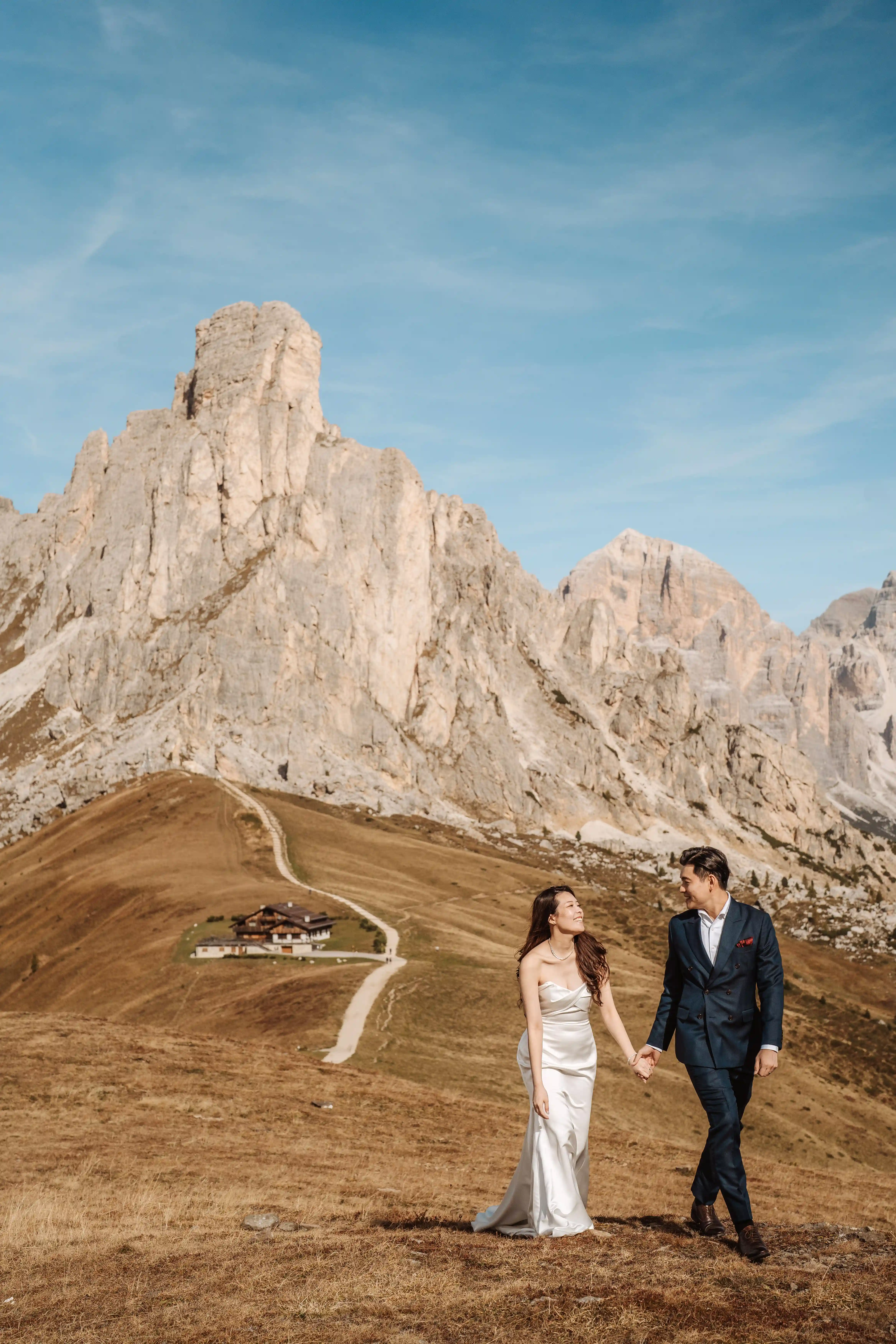Bride and groom walking hand in hand in the Dolomites with mountain peaks and alpine hut in the background