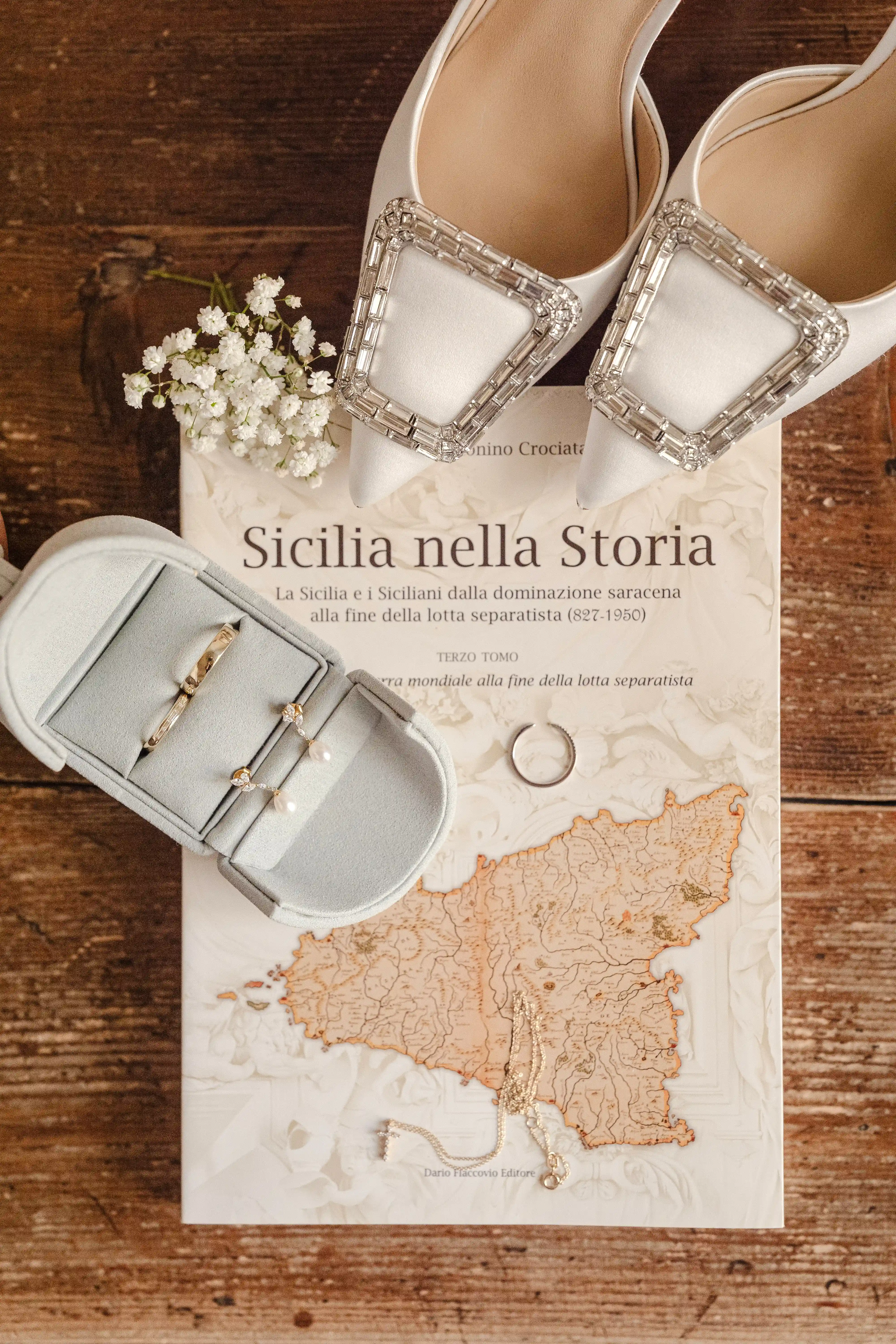 Elegant bridal shoes with jeweled buckle placed on a “Sicilia nella Storia” book with wedding rings and jewelry on a wooden table