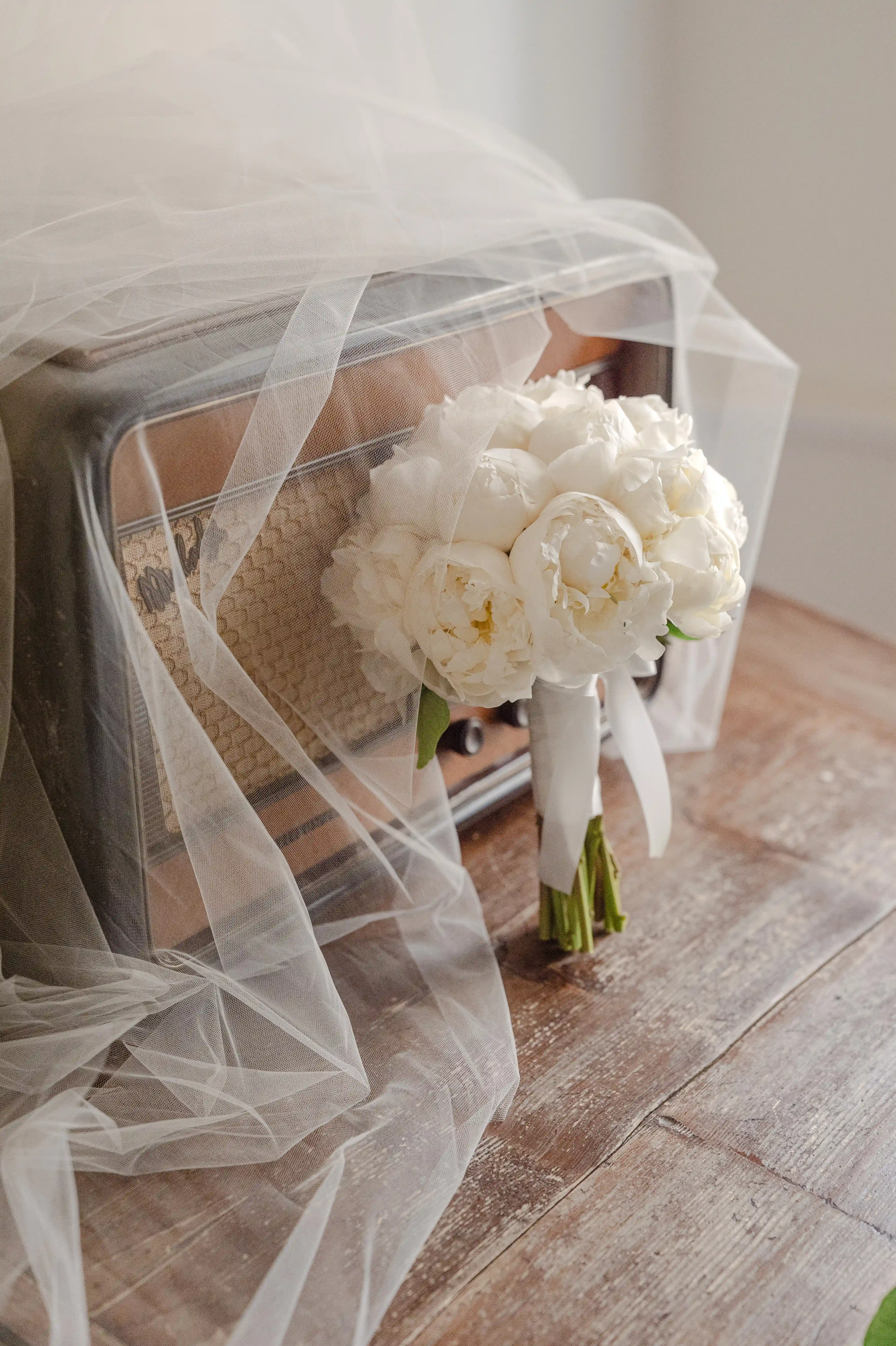 White peony bridal bouquet next to a vintage suitcase covered with a soft veil