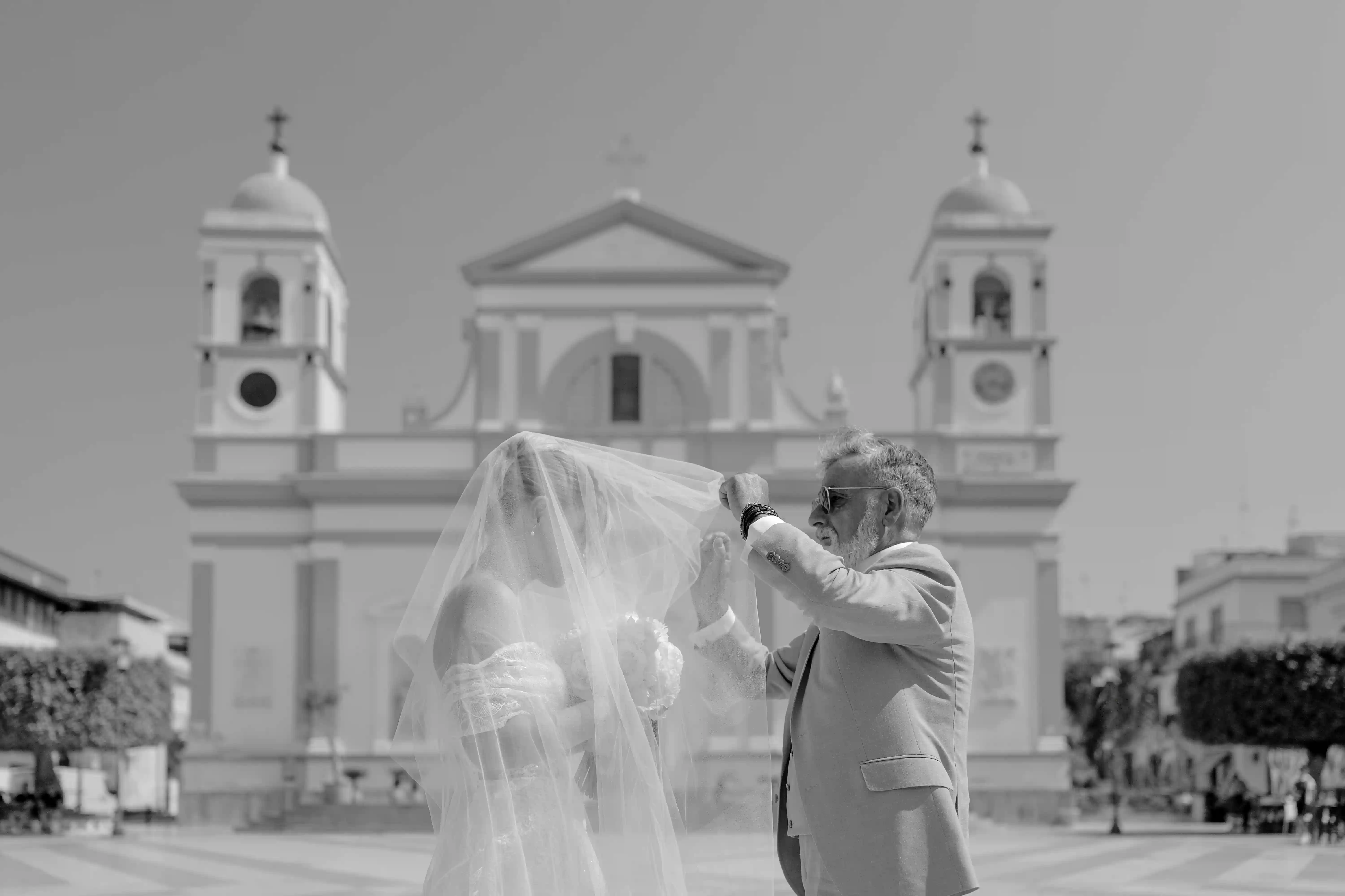 Black and white photo of a father lifting the bride’s veil in front of a Sicilian church