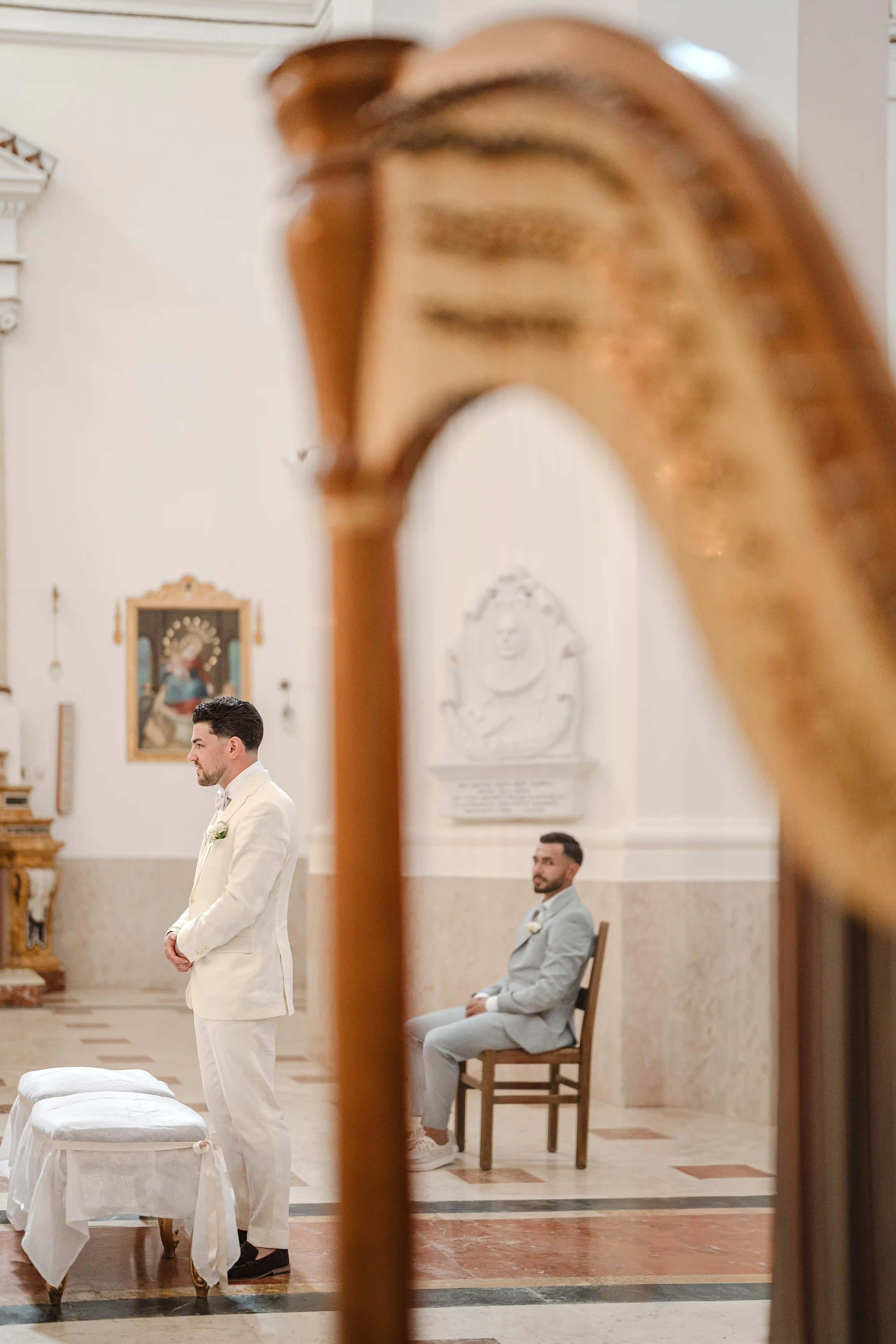 Groom seen through a mirror while standing at the altar, with a groomsman seated behind