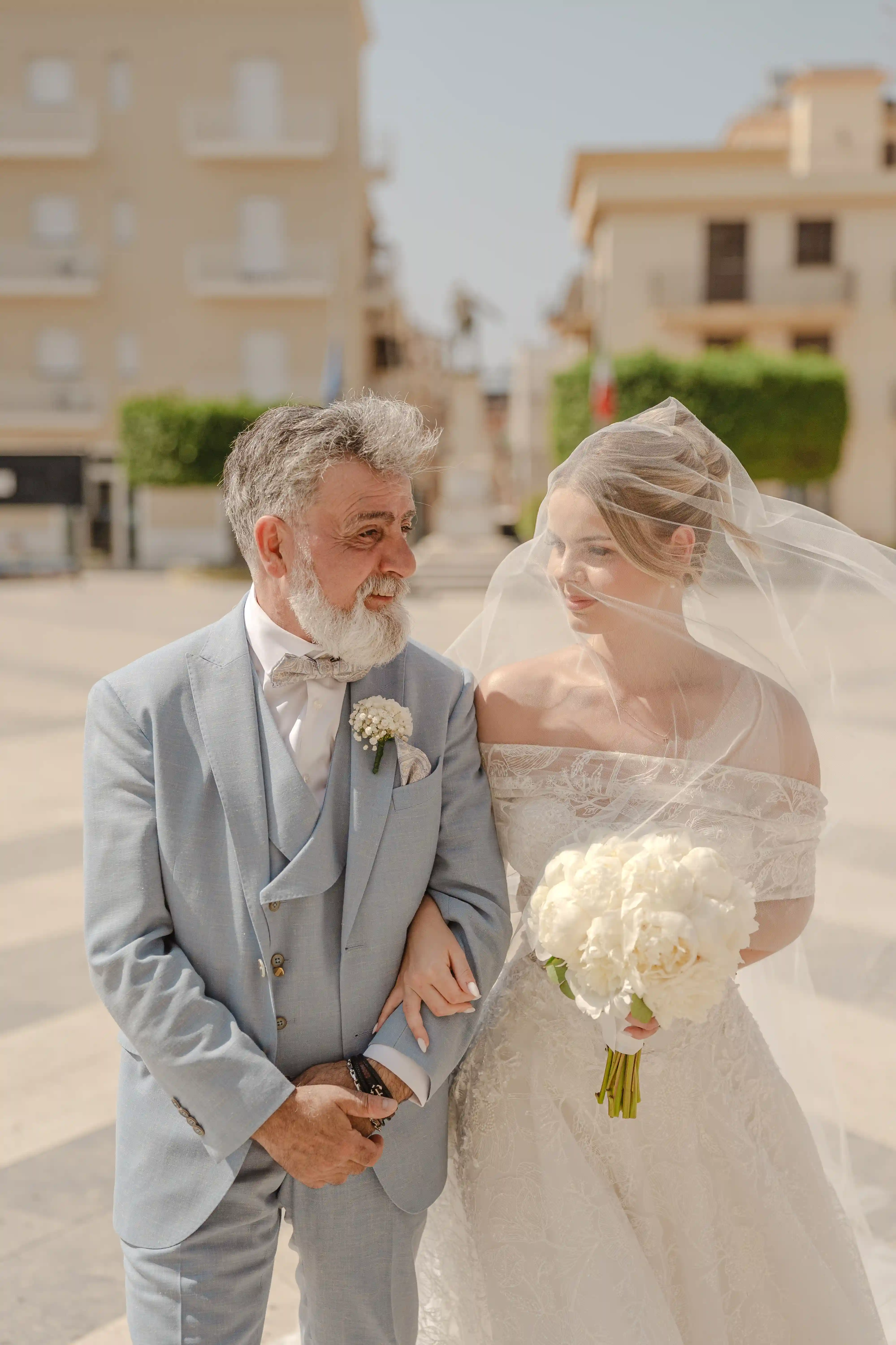 Bride walking arm in arm with her father under the veil in a sunlit Sicilian square