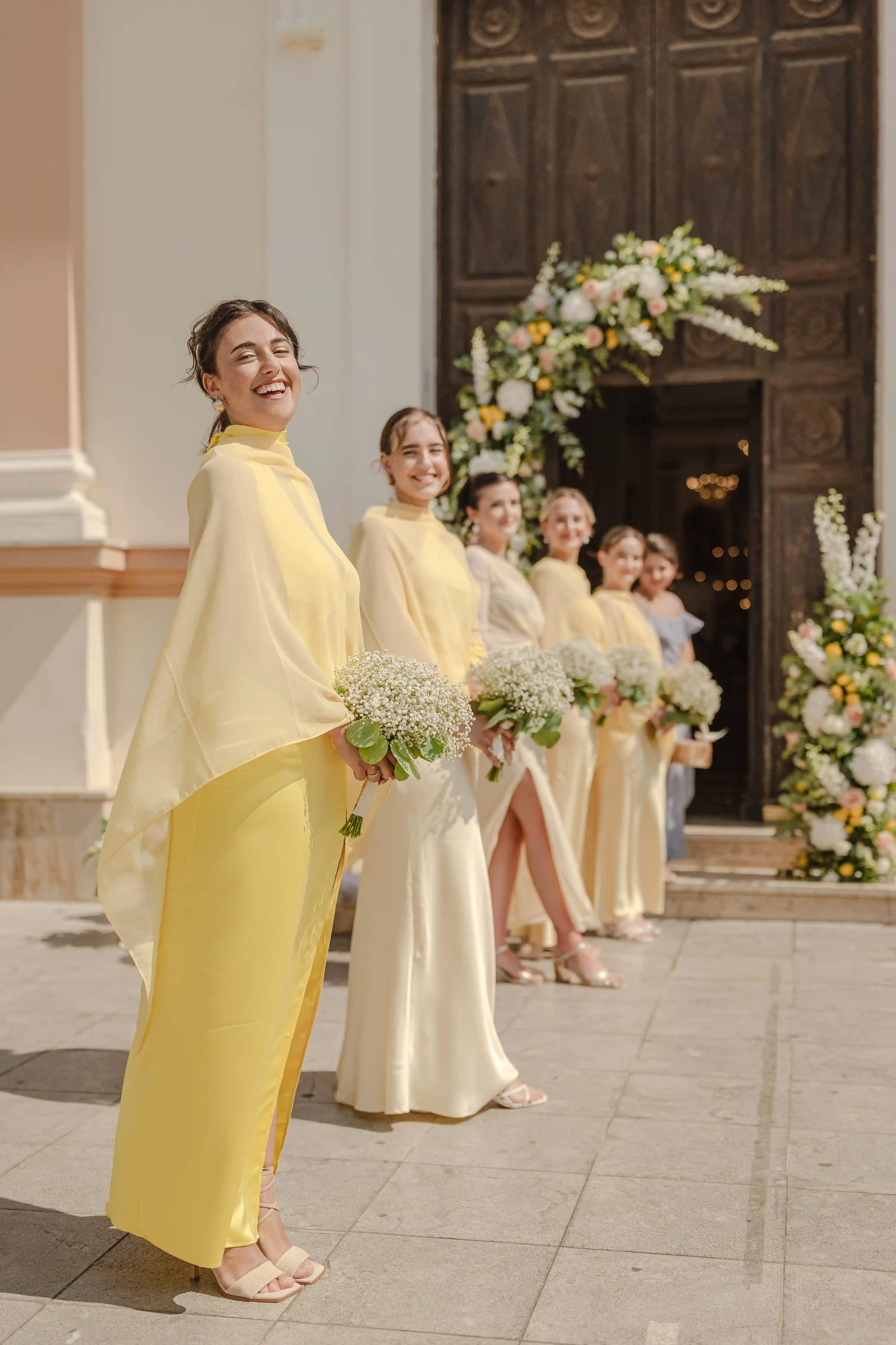 Bridesmaids in soft yellow dresses holding white bouquets outside a decorated church entrance