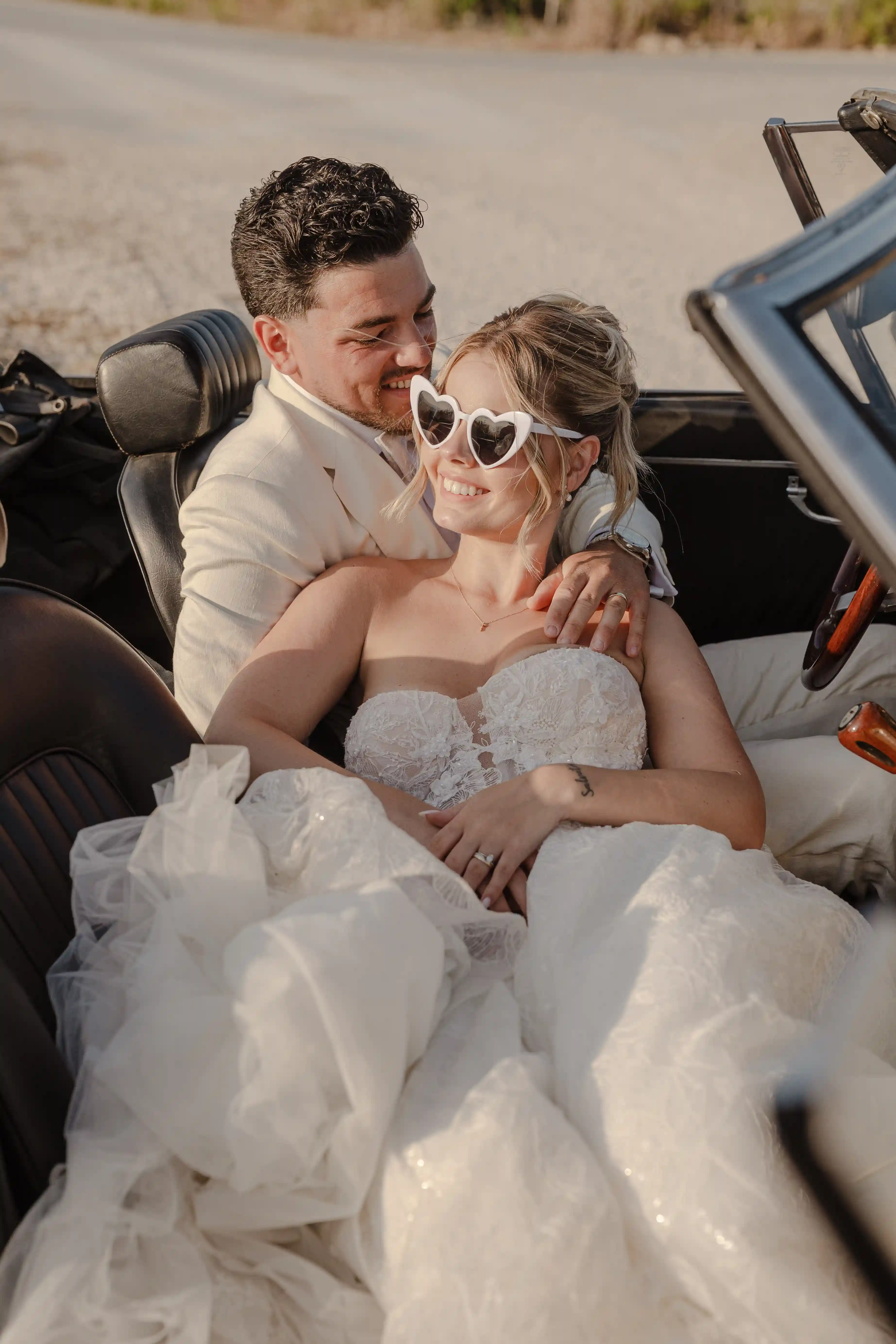 Happy bride and groom embracing in vintage convertible car at sunset