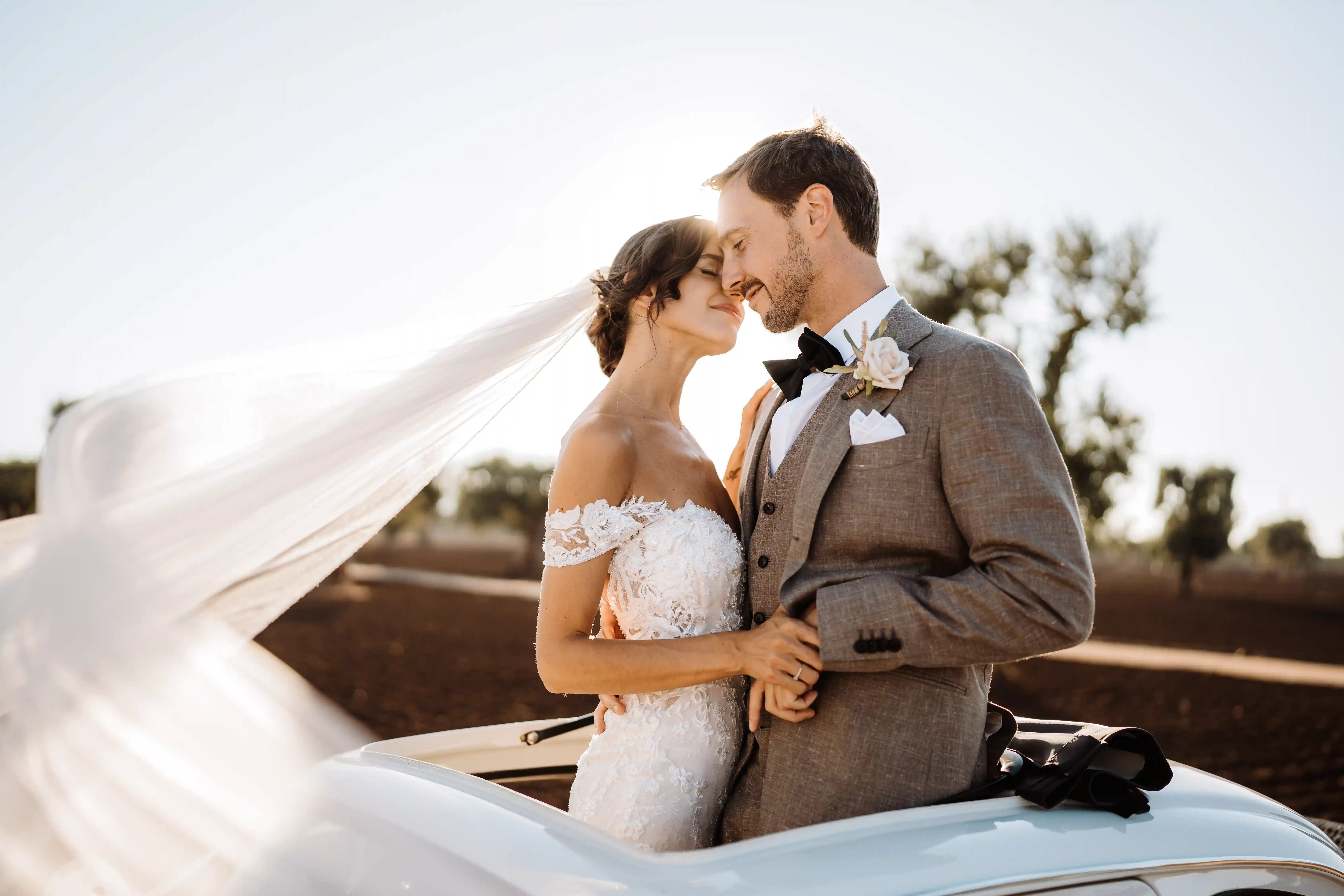 Bride and groom standing by vintage car with flowing veil in Puglia, stylish Italian countryside wedding