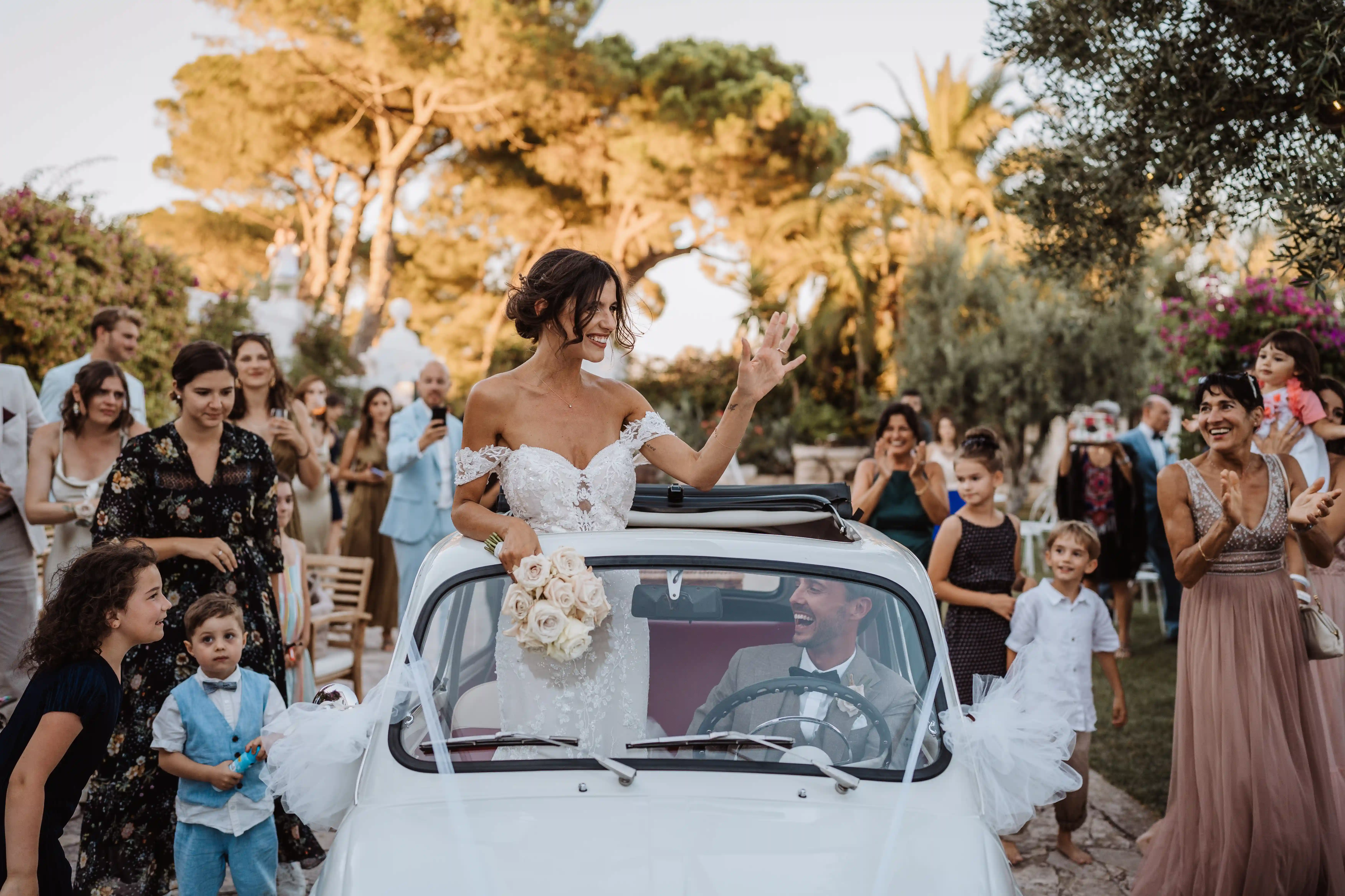 Bride standing in a classic Fiat 500 waving to guests during outdoor wedding reception in Italy