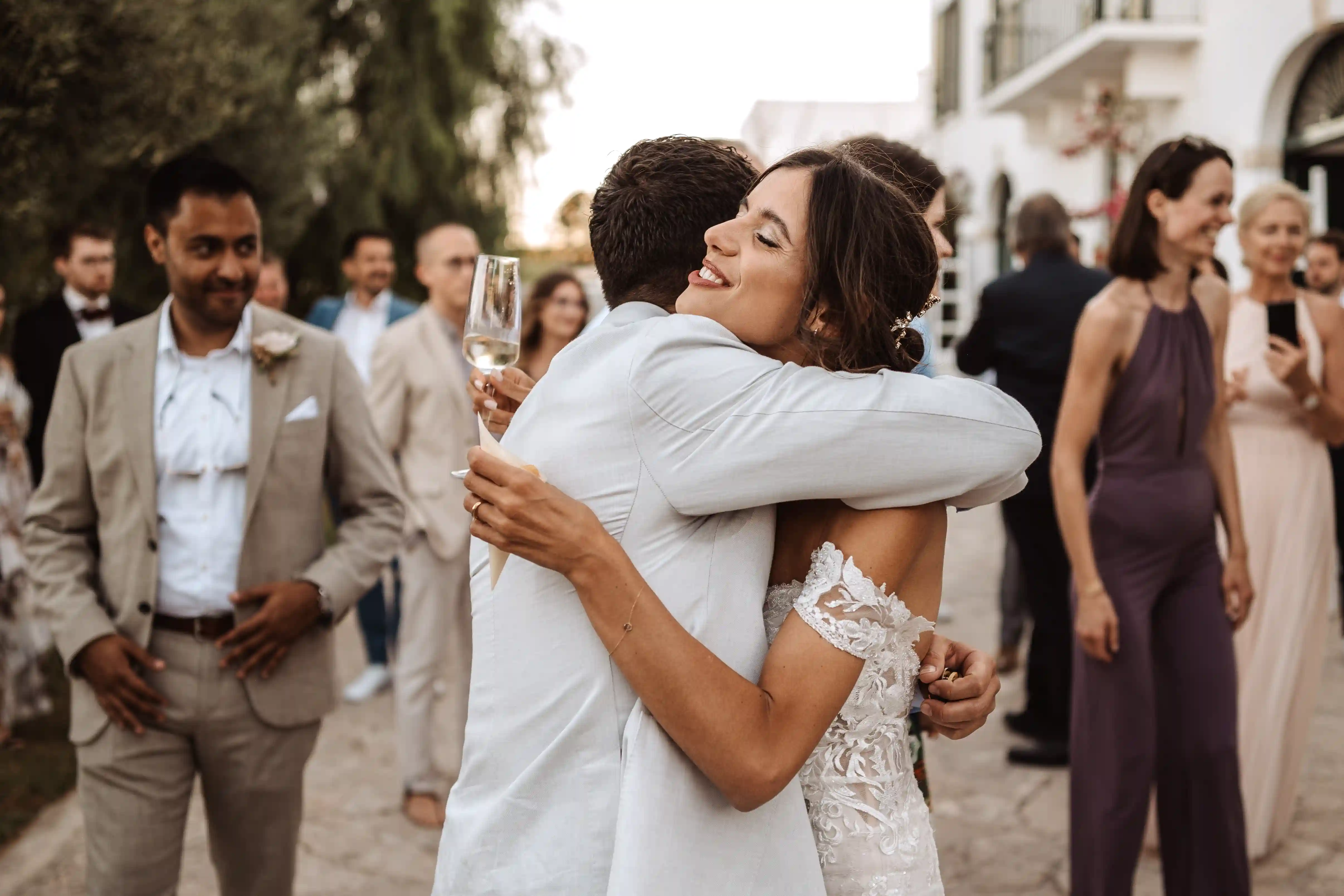 Bride hugging a guest while holding a glass of champagne at a summer wedding reception