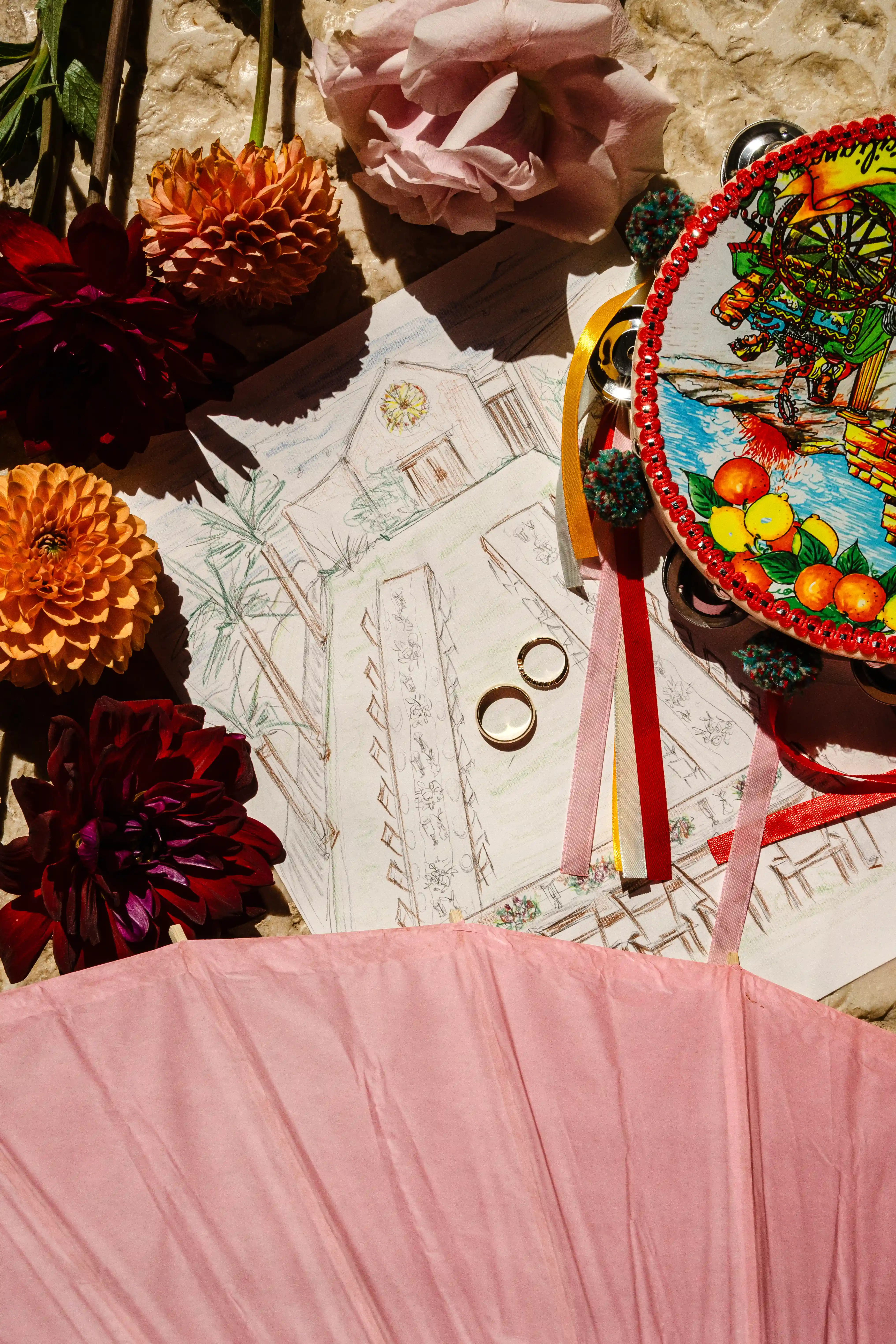 Colorful Sicilian-inspired wedding details with flowers, tambourine, and rings arranged on hand-drawn sketches in warm sunlight.