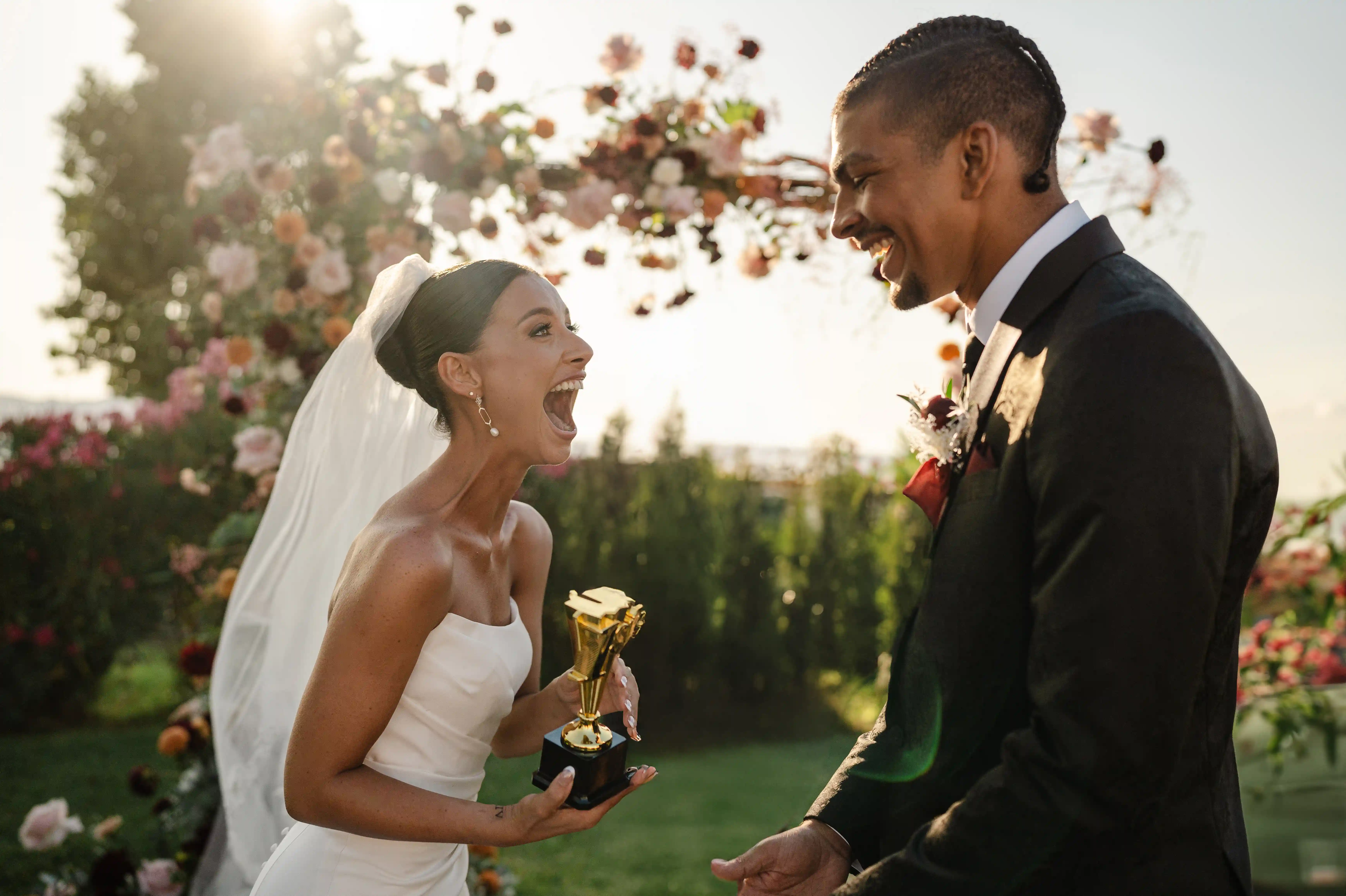 Bride in a strapless wedding dress laughing joyfully while holding a small golden trophy, facing the groom during an outdoor ceremony at sunset with floral arch in the background.