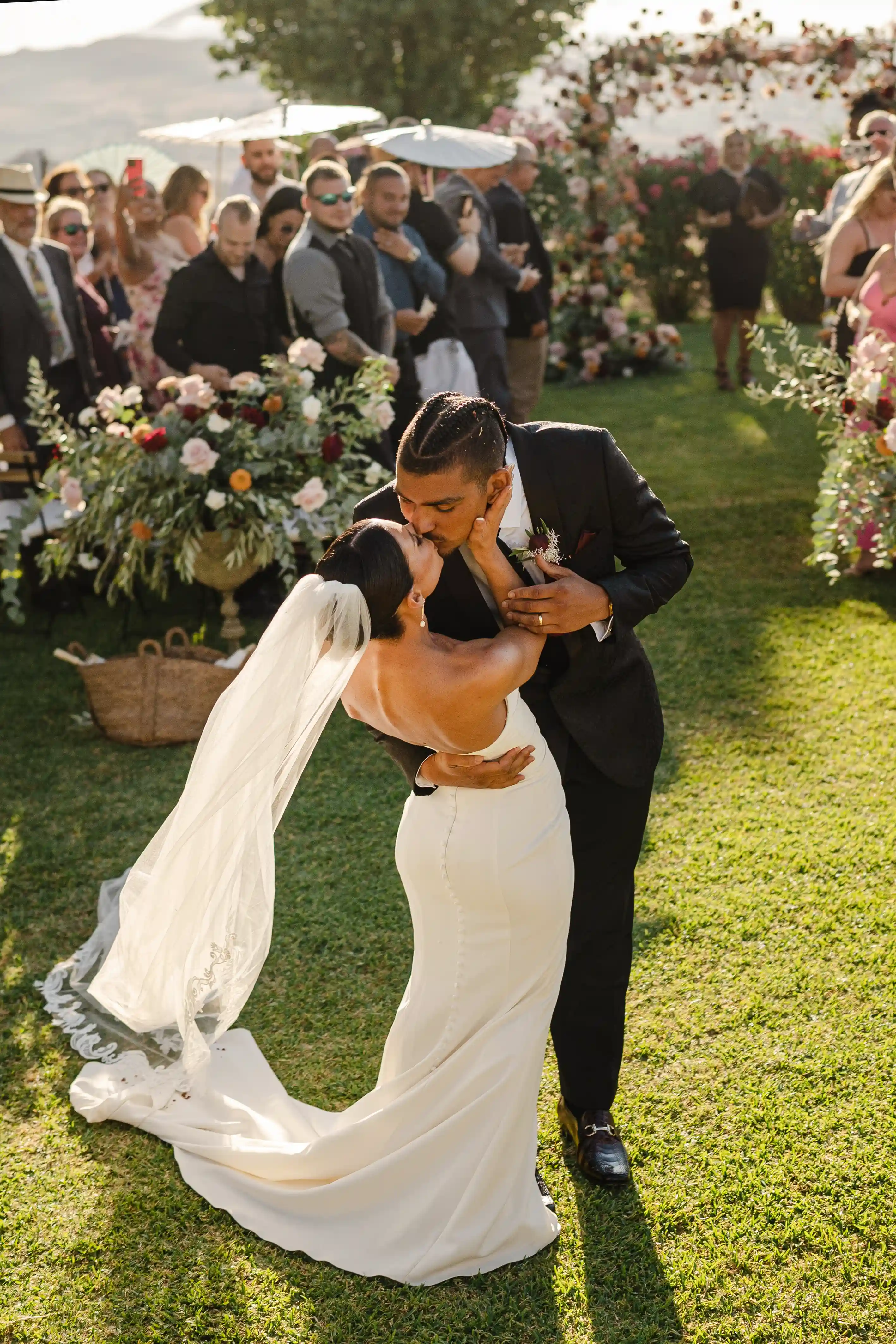 Groom dipping the bride for a romantic first kiss during an outdoor wedding ceremony, surrounded by guests and floral arrangements under warm sunlight.