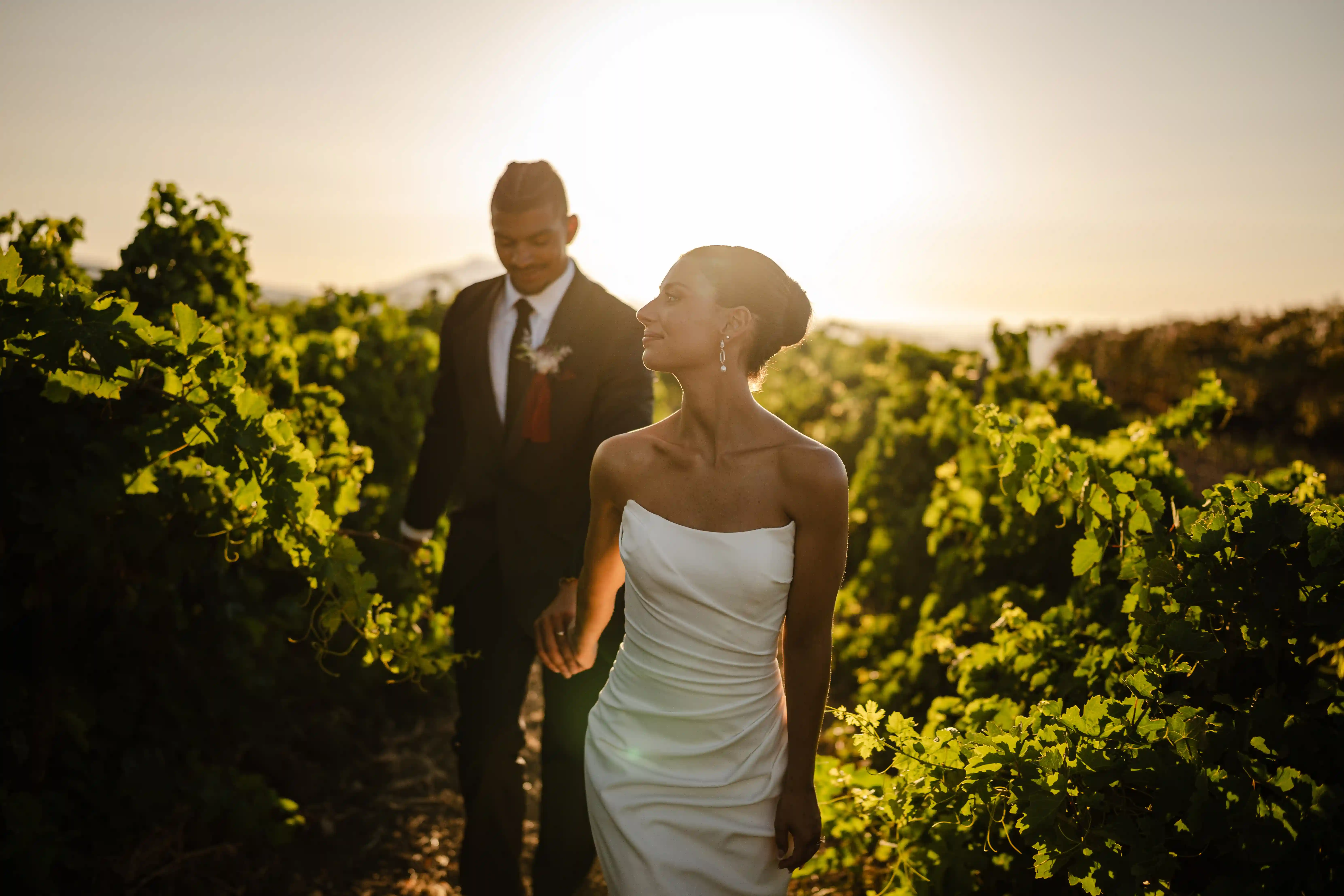 Bride and groom walking hand in hand through a vineyard at golden hour, backlit by warm sunlight creating a romantic and intimate atmosphere