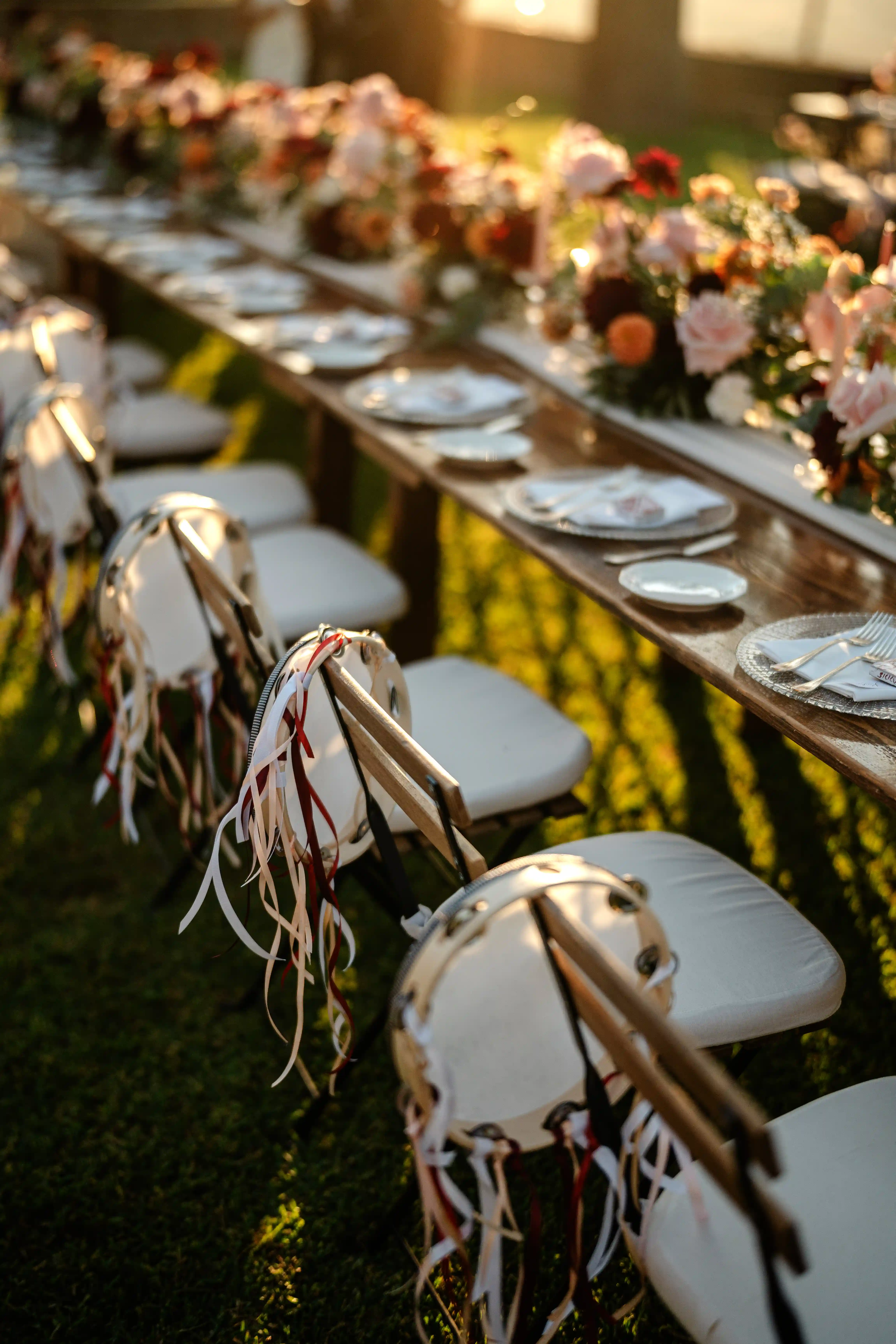 Long outdoor wedding reception table decorated with floral centerpieces, elegant table settings, and chairs adorned with ribbons in soft sunset light